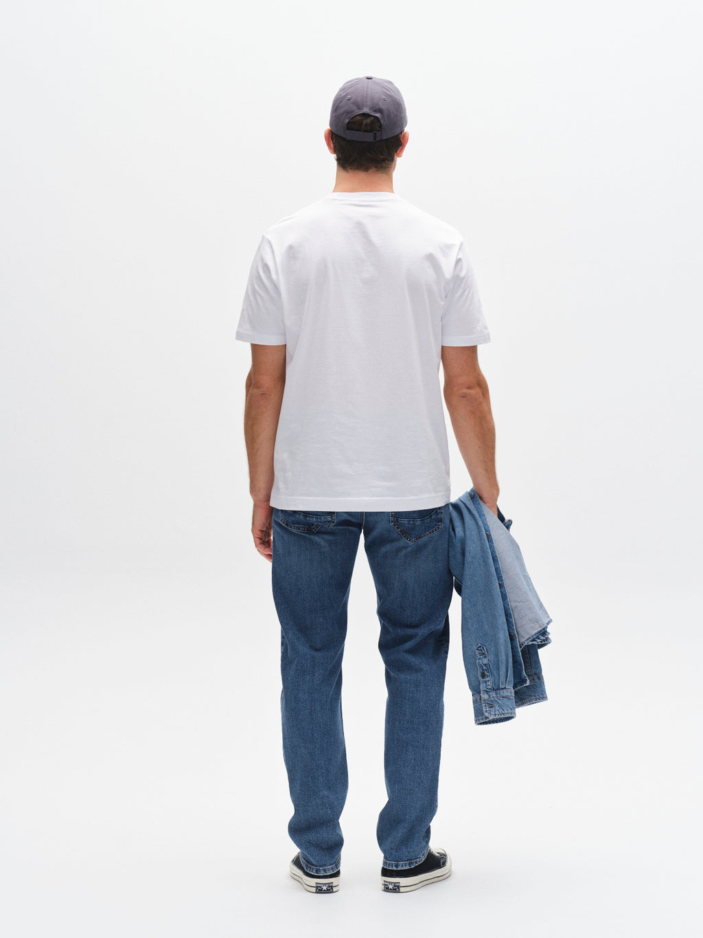 A man faces away from the camera, wearing a gray cap, white T-shirt, regular fit jeans, and sneakers. He holds a Gabba GABmarc F1010 - Dark Blue Denim jacket in his left hand against a plain white background.