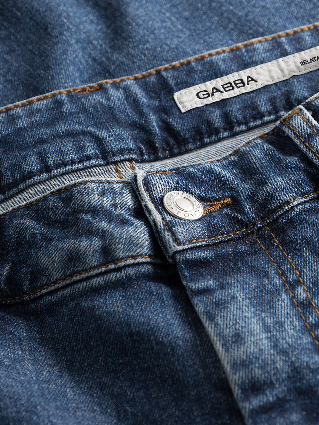 Close-up of GABmarc F1010 dark blue denim jeans by Gabba, featuring a metallic button, orange stitching, belt loops, and a white fabric label with the Gabba brand name sewn onto the waistband.