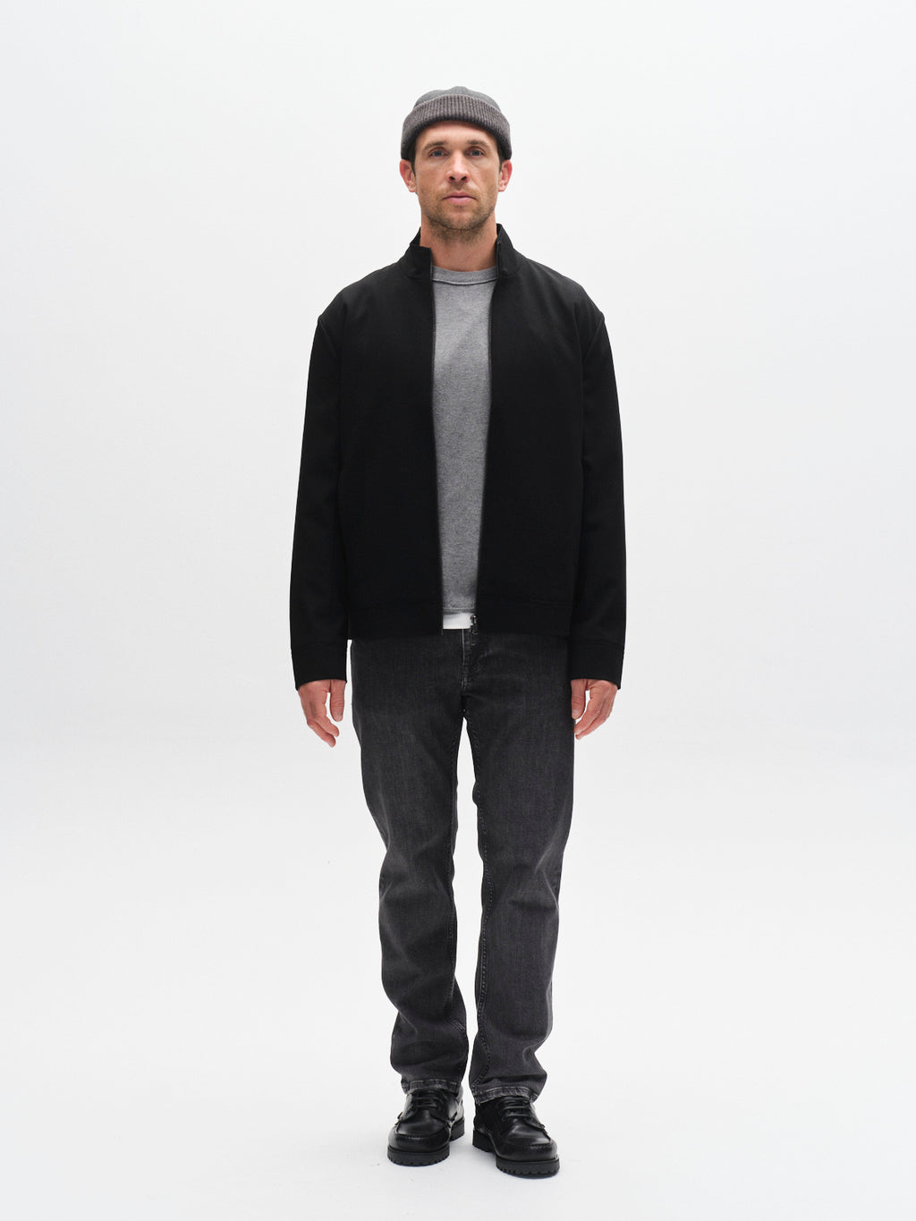 A man faces forward against a plain white background, wearing a gray beanie, black jacket over a gray shirt, Gabba GABmarc F1011 - Black Denim jeans, and black shoes.