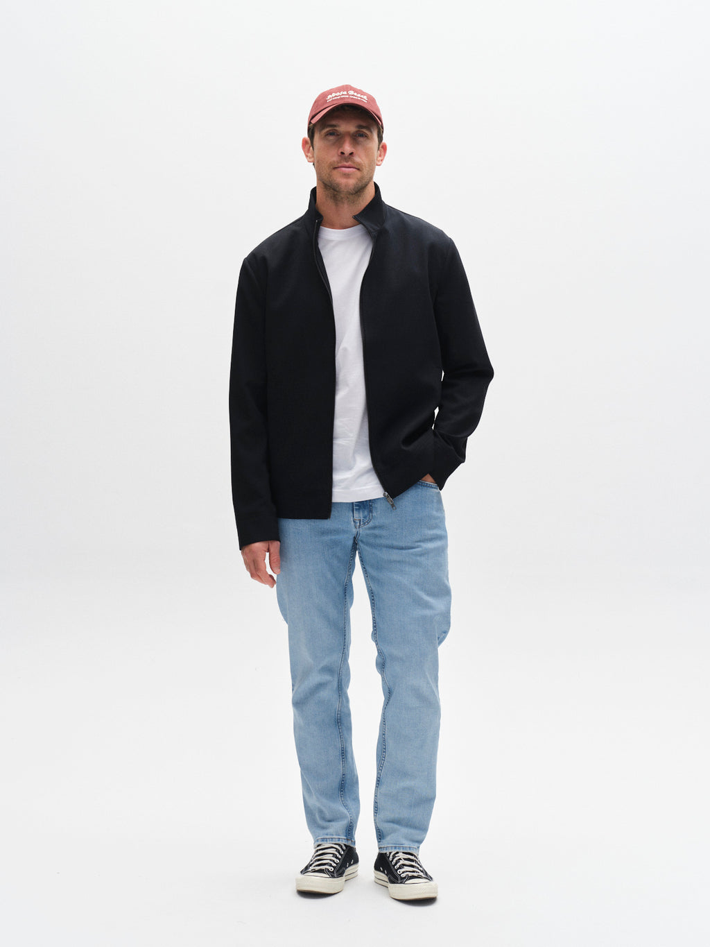A man in a red cap, black jacket, white t-shirt, Gabba GABmarc F1012 light blue denim jeans, and black sneakers stands against a plain white background.