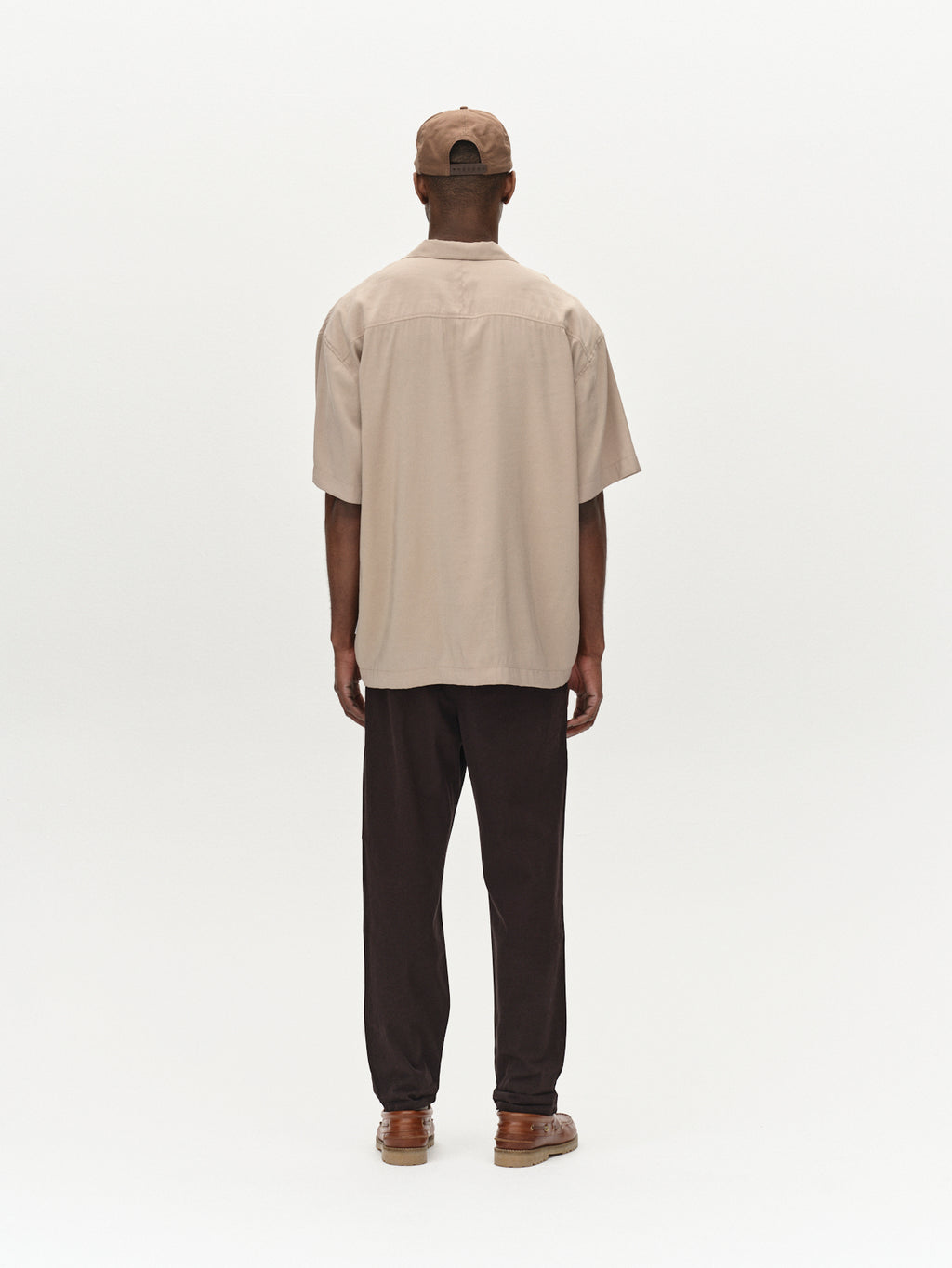 A person stands facing away, wearing Gabbas Firenze Lubo - Black chinos, beige short-sleeve shirt, brown shoes, and a beige cap against a plain white background.