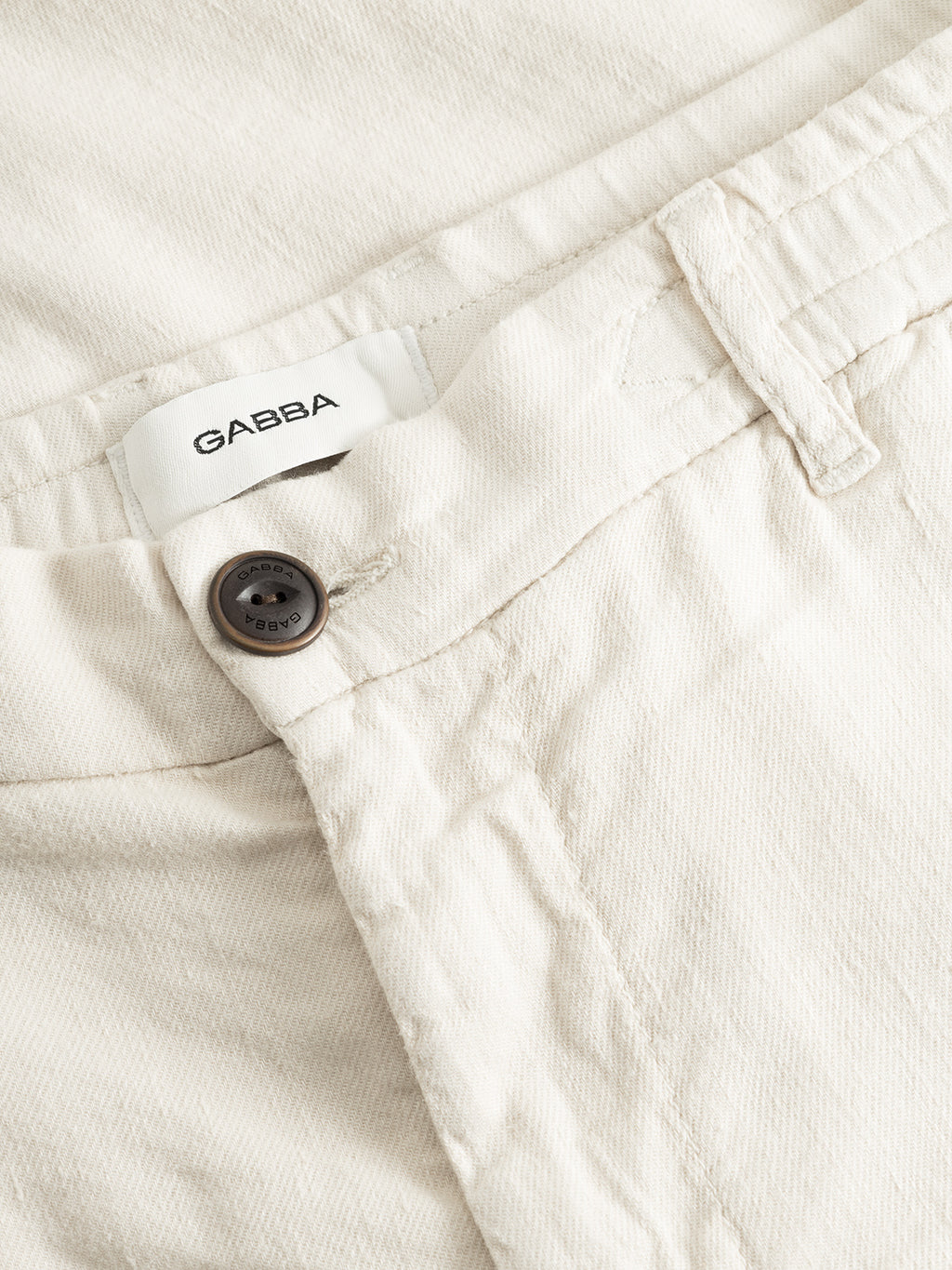 Close-up of the waistband and button of Gabba Monza Twill Linen - Birch pants, featuring a subtle textured fabric, brand label, and a single dark button above the zipper—ideal for a relaxed straight fit.