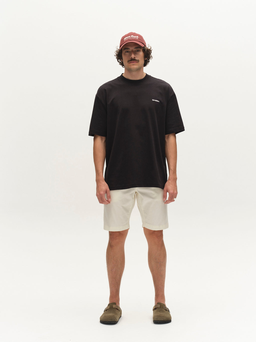 A man faces forward against a plain white background, wearing a maroon cap, black t-shirt, Gabba GABjet K3280 Dale Shorts in white, and brown slip-on shoes—an ideal everyday look.
