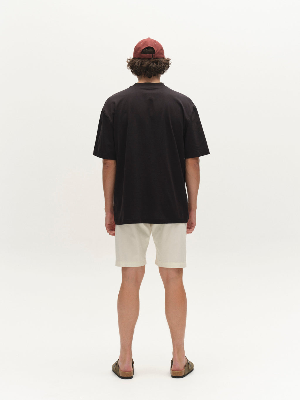 A person stands facing away, wearing black T-shirt, Gabba GABjet K3280 Dale Shorts in white, olive sandals, and a red baseball cap against a plain white background.