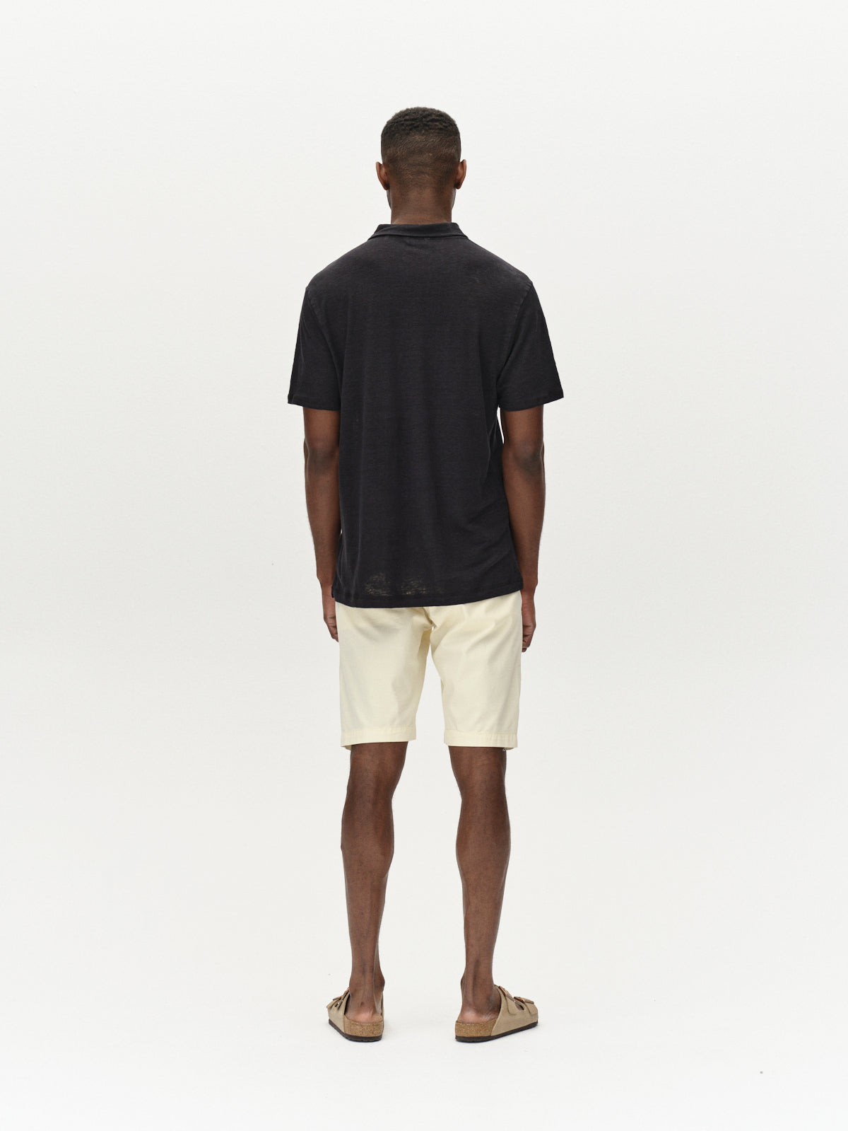 A man stands facing away against a white background, wearing a black short-sleeve shirt, Jet K3280 Dale Shorts - Ecru Light from Gabba, and brown sandals. The comfortable fit exemplifies versatile design, perfect for any casual occasion.