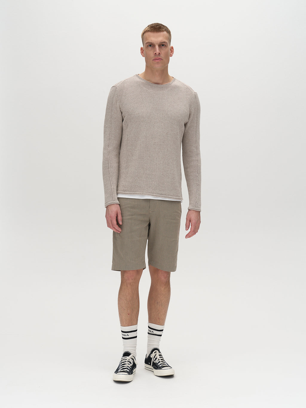 A man stands in a neutral studio, wearing a beige long-sleeve shirt, the Gabba GABjet K3280 Dale Shorts in Humus with a comfortable fit, striped socks, and black sneakers—a look defined by its timeless design.