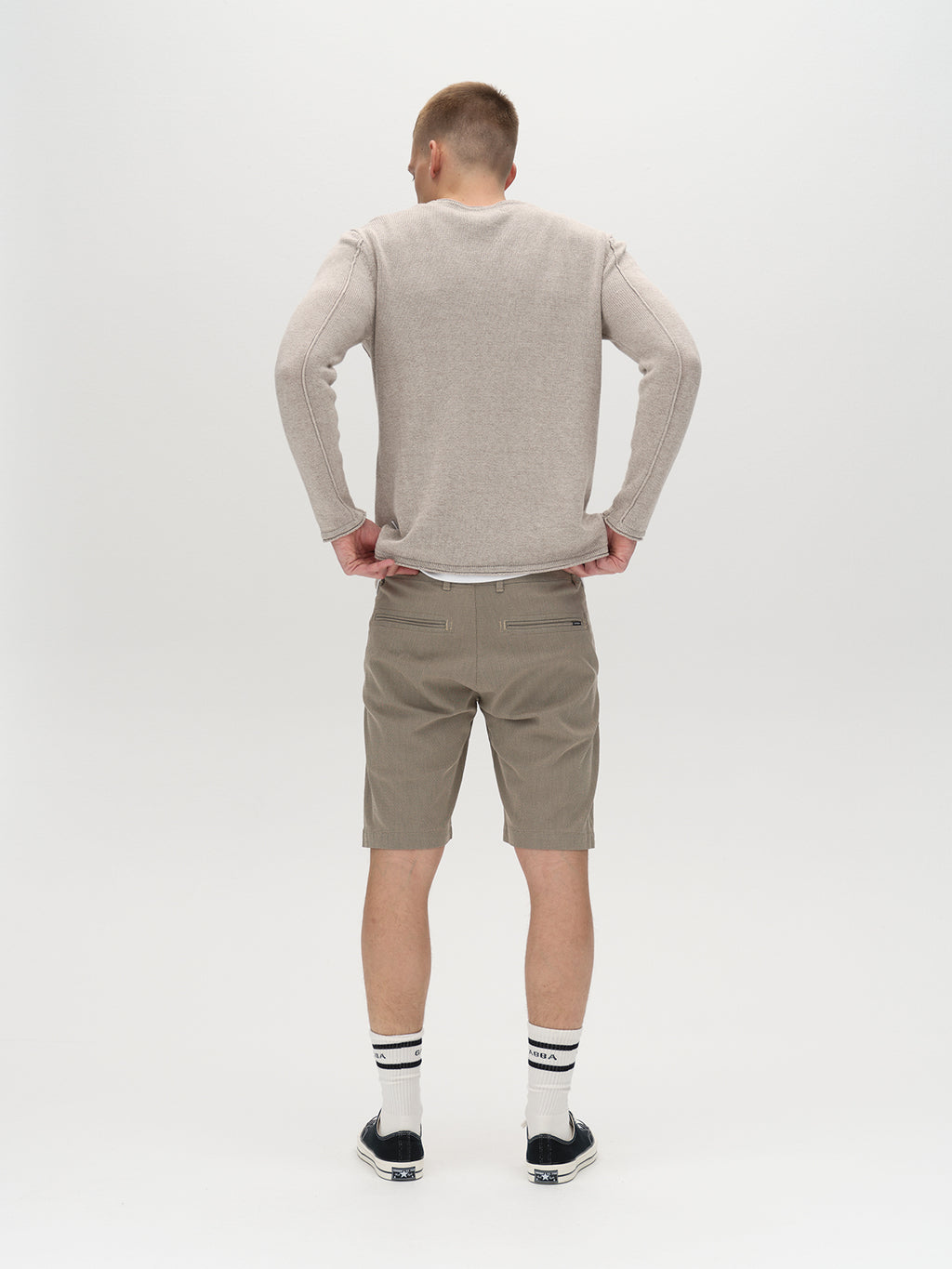 A man stands with his back to the camera, wearing a light beige long-sleeve shirt paired with Gabba’s GABjet K3280 Dale Shorts in Humus, striped socks, and black sneakers against a plain white background. The ensemble highlights both comfortable fit and timeless design.