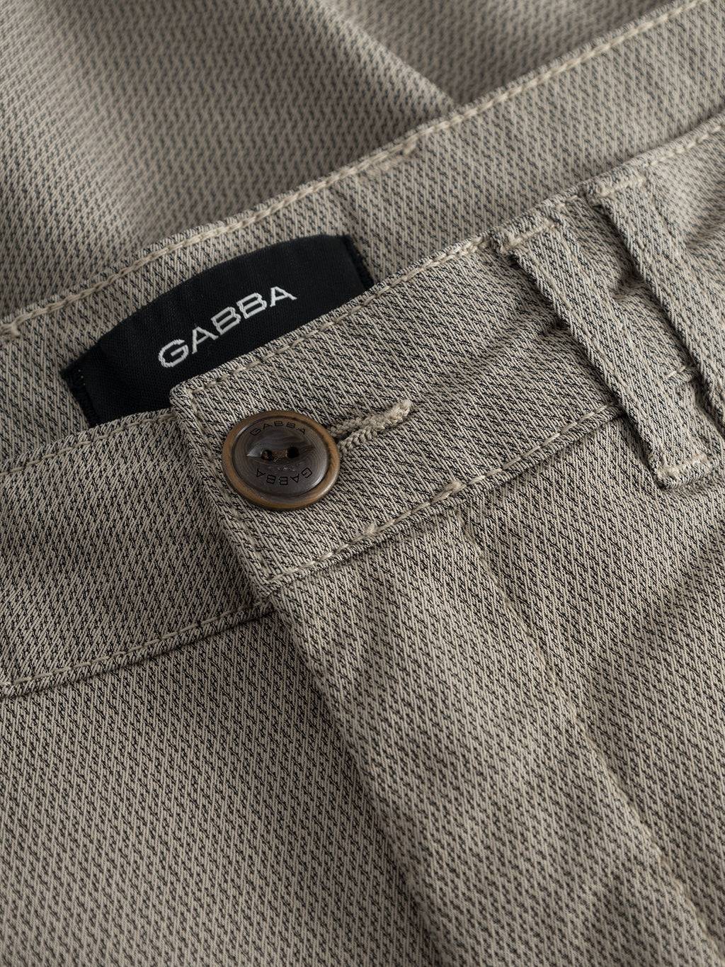 Close-up of the GABjet K3280 Dale Shorts in Humus by Gabba, featuring a beige textured fabric with a metal button, waistband, belt loop, and a visible black fabric label reading “GABBA.” These shorts offer a timeless design and are crafted for comfortable wear.