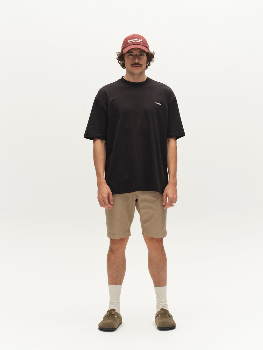 A man with a mustache and neutral expression stands against a white background wearing a black T-shirt, white socks, brown sandals, a red baseball cap, and Gabba GABjet K3280 Dale Shorts in Sand.