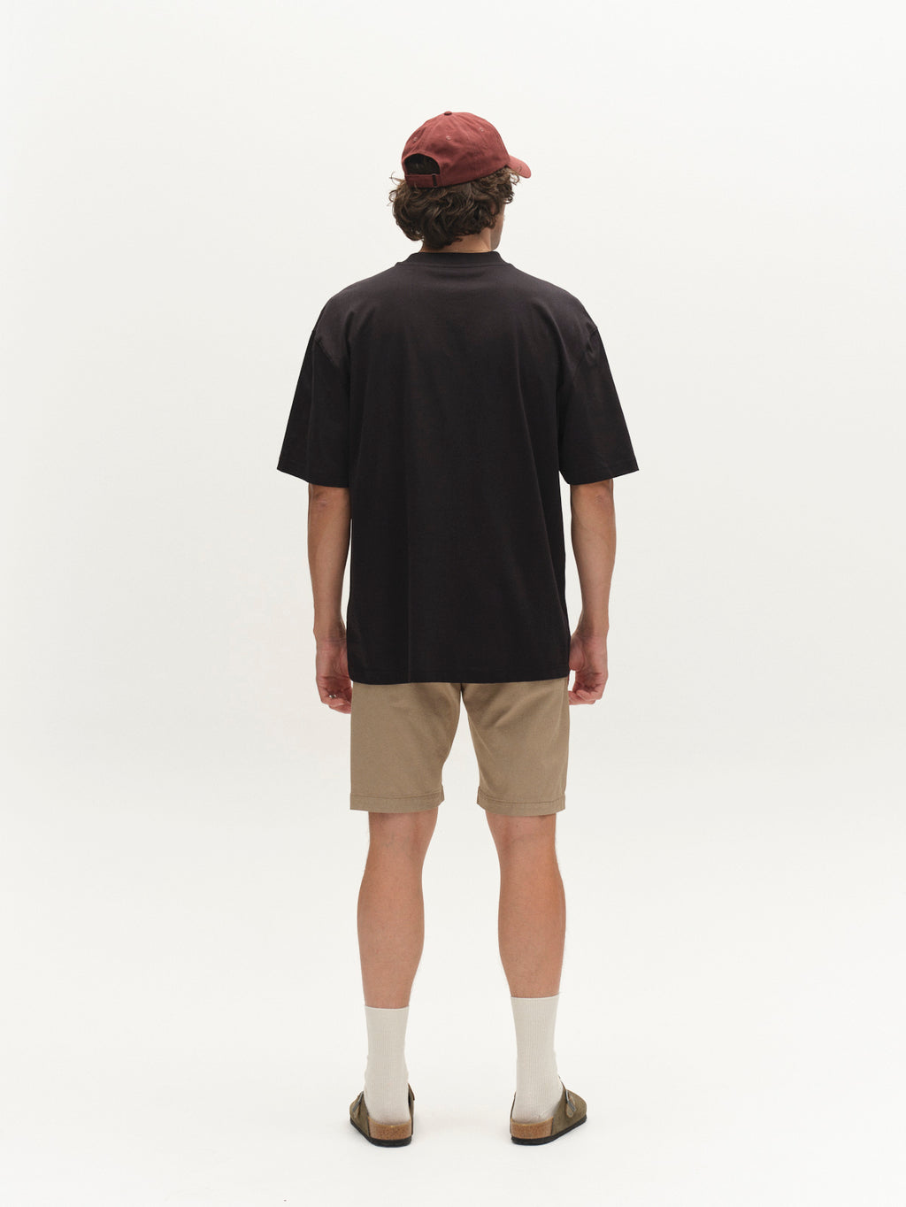 A person faces away, wearing a black T-shirt, Gabba GABjet K3280 Dale Shorts in Sand, white socks, brown sandals, and a red cap against a plain white background.