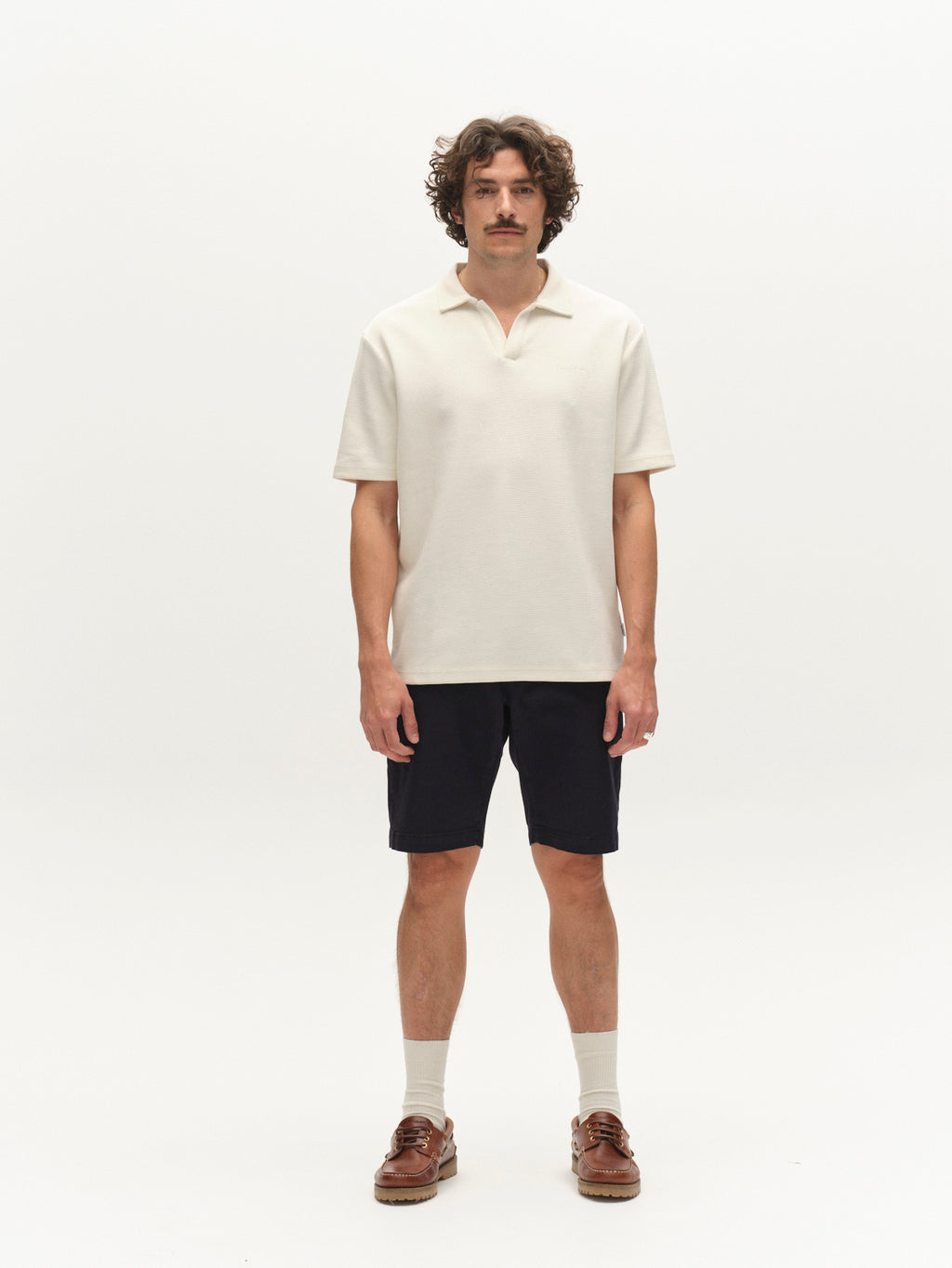 A man with curly hair and a mustache stands on a white background wearing a cream polo shirt, Gabba GABjet K3280 Dale Shorts in Blue Black, white socks, and brown loafers—a timeless, effortless look.