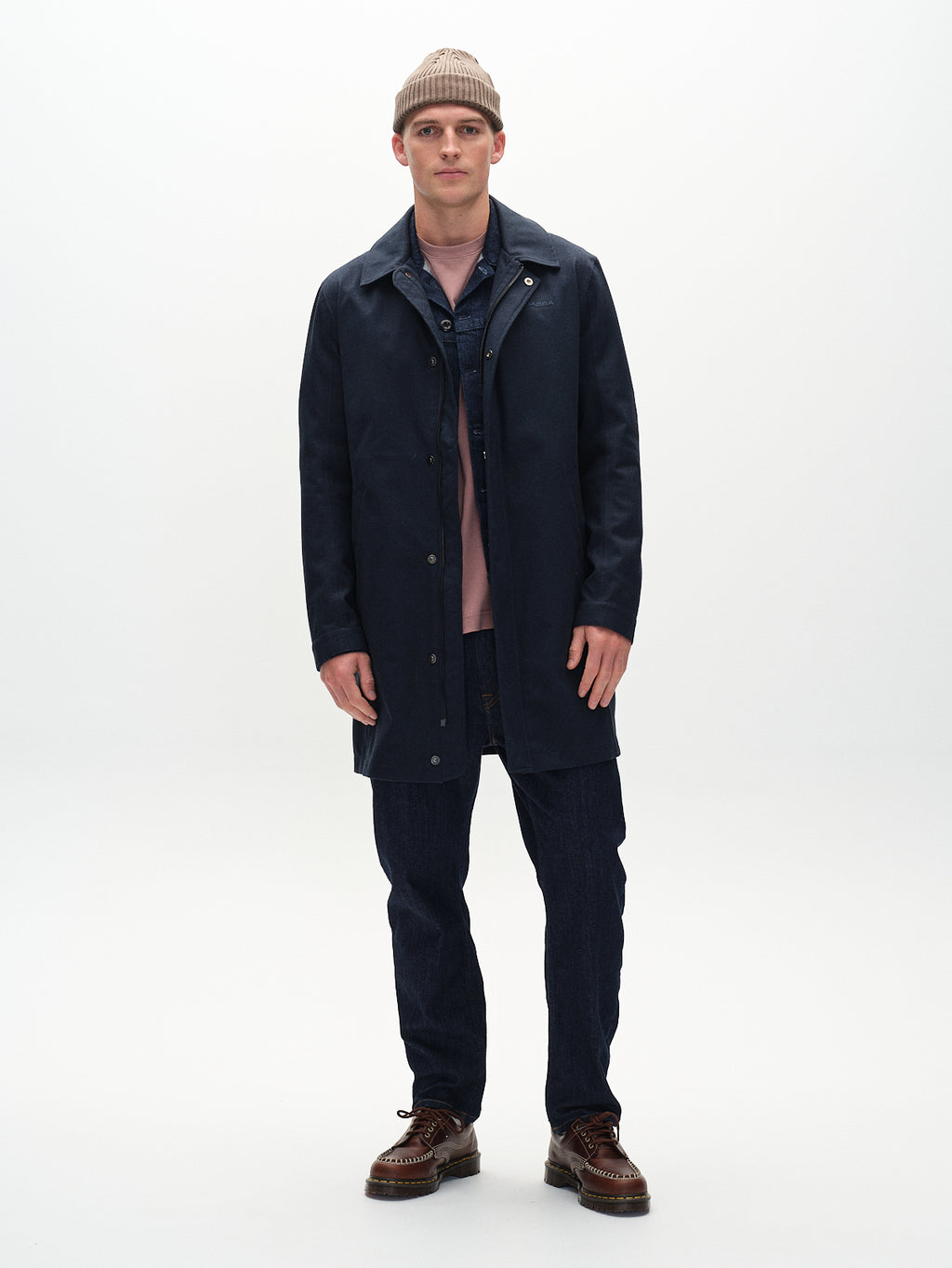 A young man stands facing forward, wearing a tan beanie and the Gabba Emerson Bru - Navy jacket—a wind-resistant style with a TPU membrane—over a denim jacket and pink shirt, paired with dark jeans and brown shoes, set against a plain white background.
