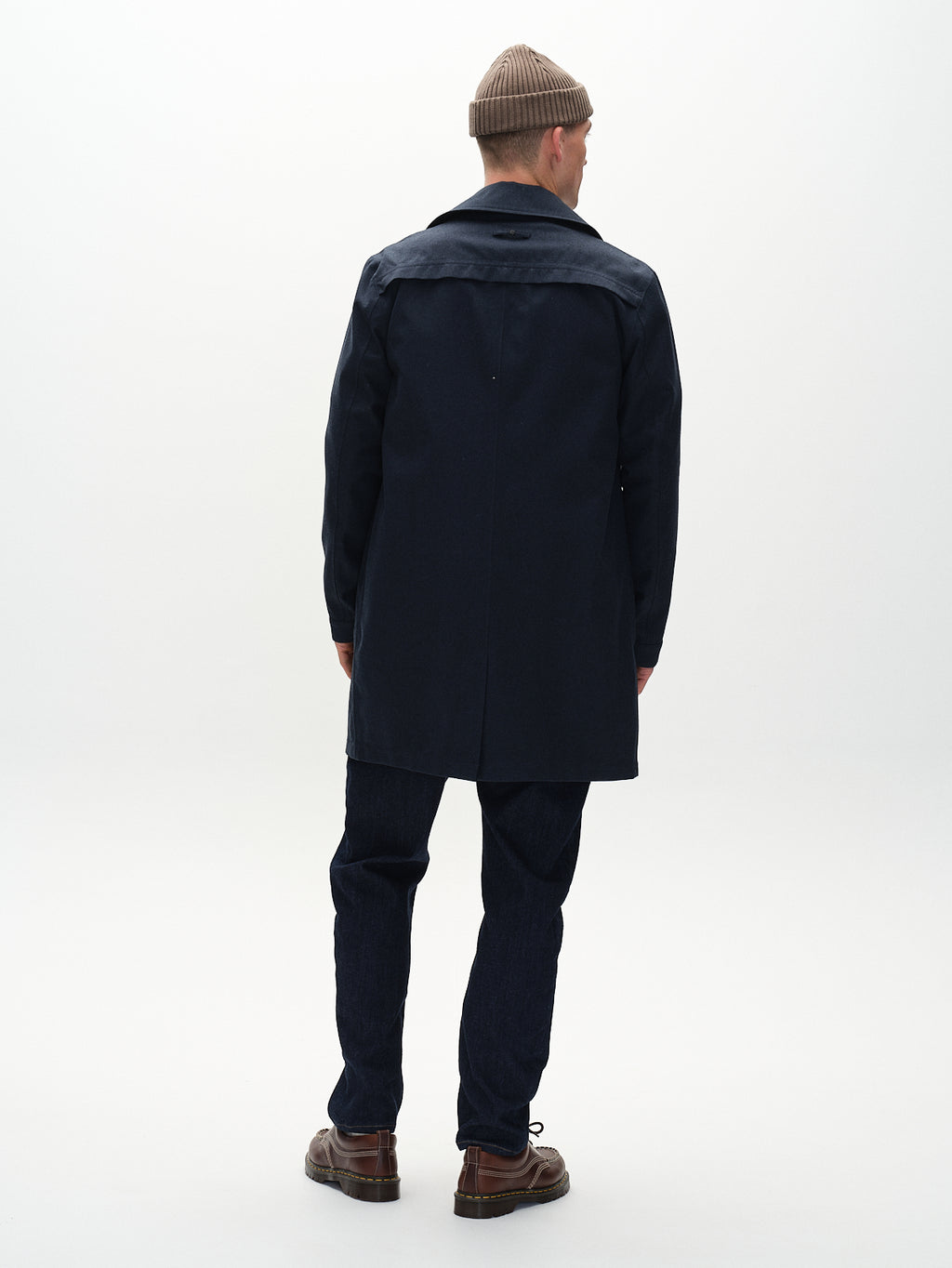 A person wearing a beige knit beanie, Gabba Emerson Bru - Navy jacket with wind-resistant TPU membrane, dark jeans, and brown shoes stands facing away from the camera against a plain white background.