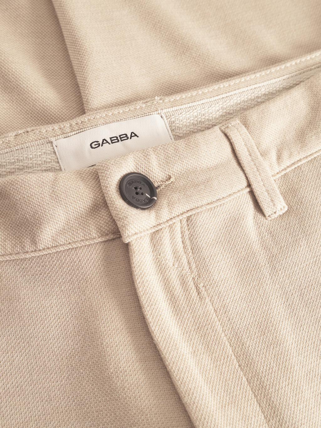 Close-up of Paul GKnit - Sand Plaza pants by Gabba, featuring a black button, belt loop, and white Gabba label inside the waistband. The textured woven fabric offers a regular slim fit, ideal for everyday wear.