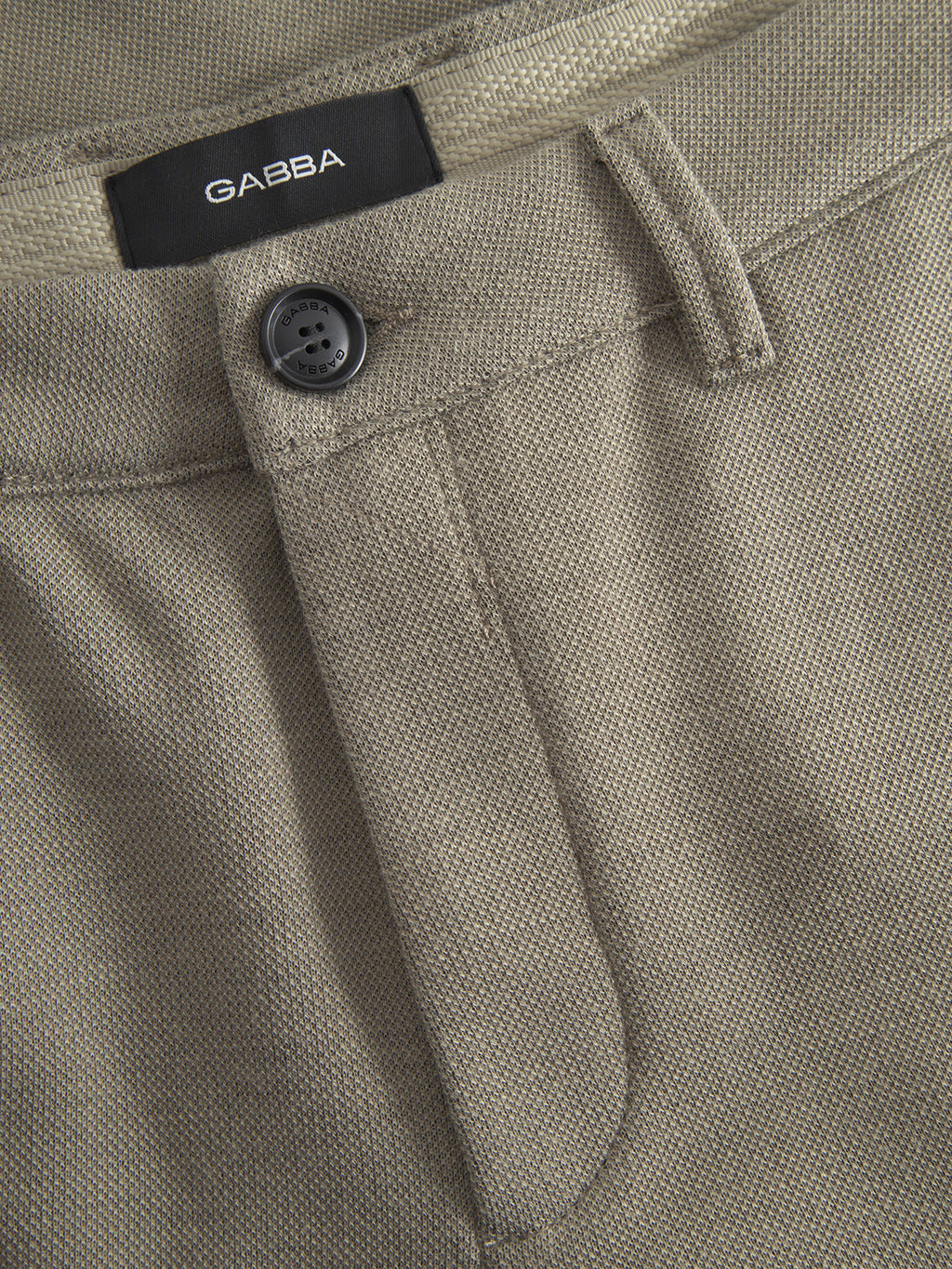 Close-up of light green Paul GKnit slim fit pants by Gabba, featuring a black button, belt loop, and a black Gabba label on the waistband. The textured structure fabric adds modern style to these fashionable pants.