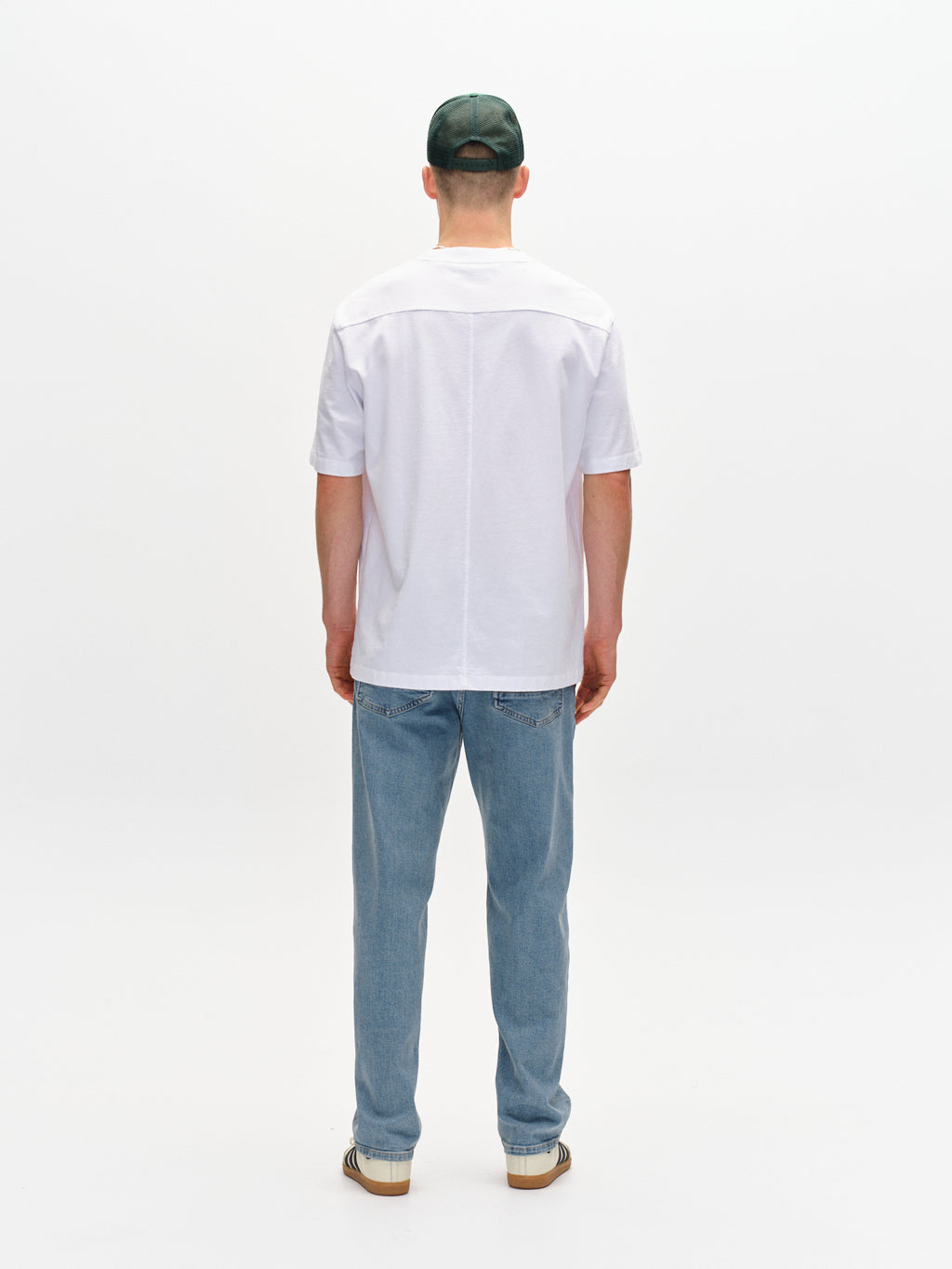 A man faces away from the camera, wearing the Gabba Nigel Wix Boxy Hour SS t-shirt in white, light blue jeans, a dark green cap, and striped shoes against a plain white background.