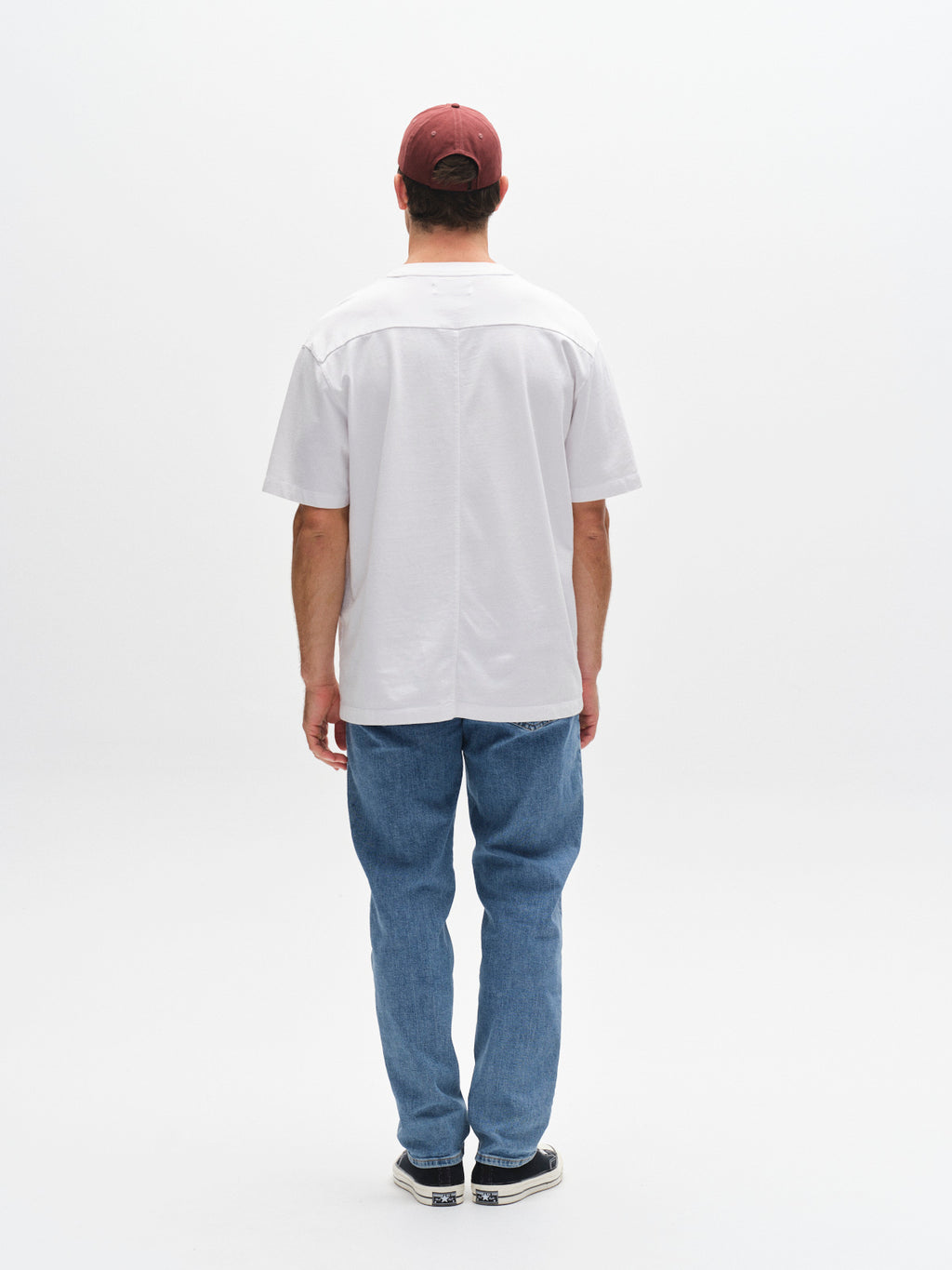 A man faces away from the camera, wearing a maroon cap, blue jeans, black sneakers, and the Gabba GABnigel Wix Boxy Hour SS tee in white against a plain white background.
