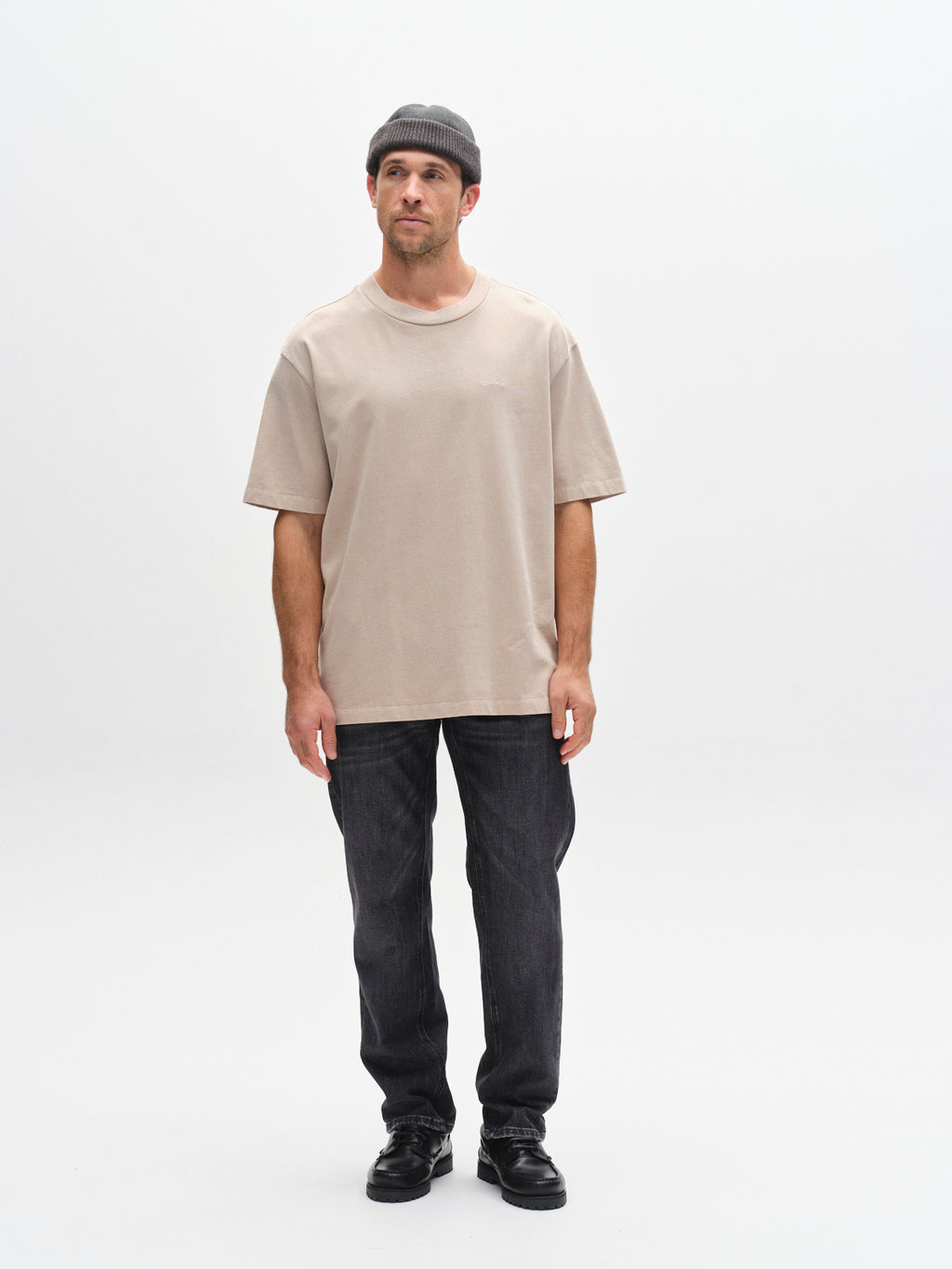 A man in a Gabba GABnigel Wix Boxy Hour SS - Sand Cashmere tee, gray beanie, black jeans, and black shoes stands against a plain white background, looking slightly left with his arms relaxed at his sides.