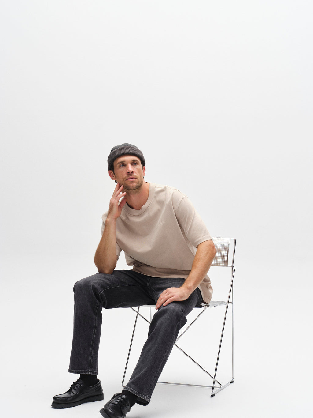 A man in the Gabba GABnigel Wix Boxy Hour SS Sand Cashmere tee, black jeans, black shoes, and a dark beanie sits on a wire chair against a plain white background, resting his chin on his hand and gazing upward thoughtfully.