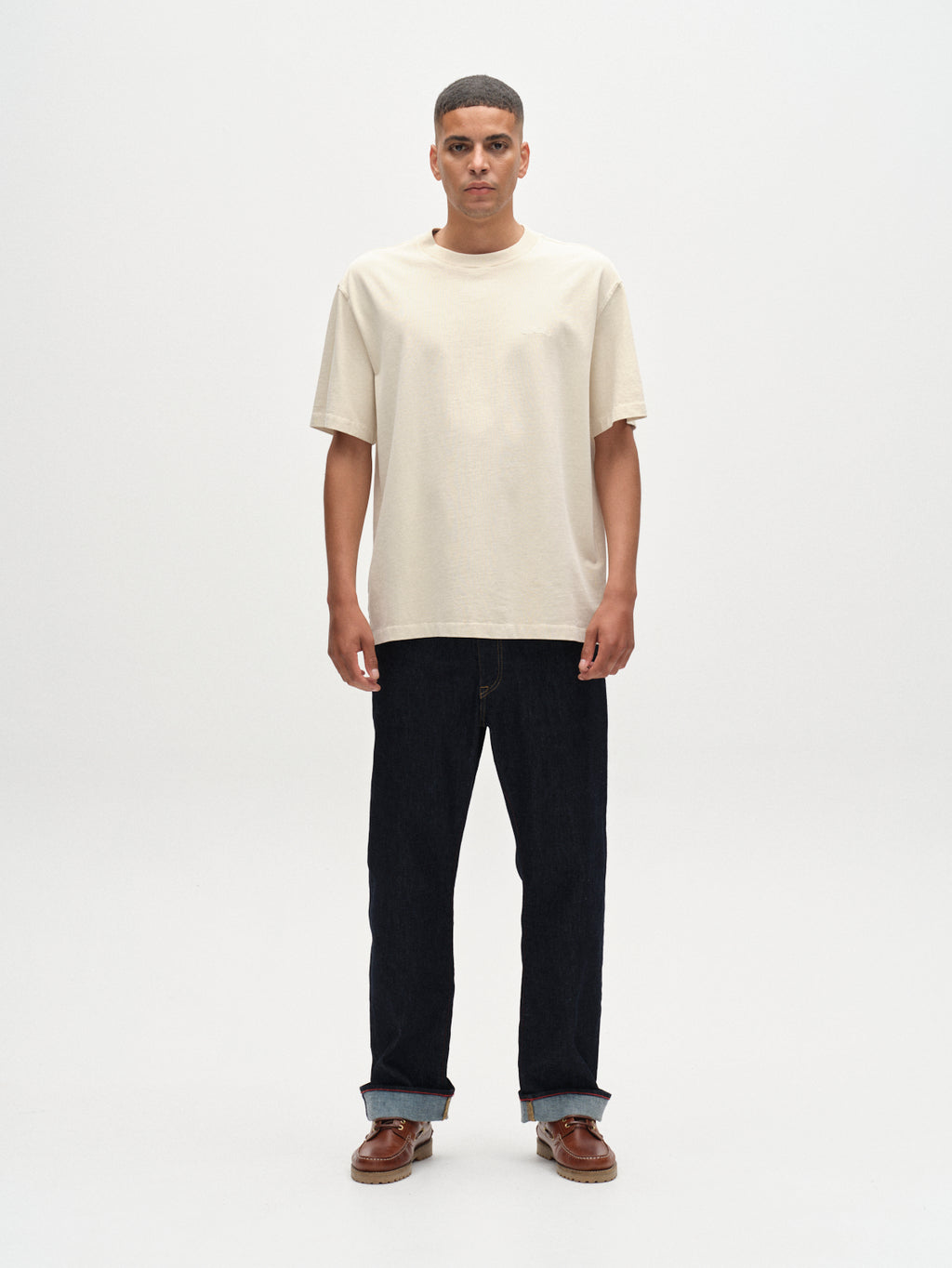 A man stands with arms at his sides, wearing the Gabba Nigel Wix Boxy Hour SS T-shirt in White Almond against a plain light background, paired with cuffed dark jeans and brown shoes. He faces forward with a neutral expression.