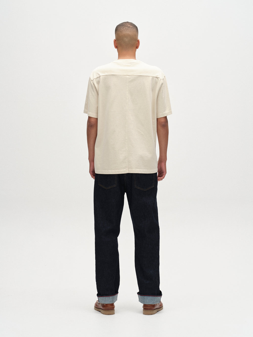 A person stands facing away from the camera, wearing the Gabba Nigel Wix Boxy Hour SS T-shirt in White Almond, paired with dark jeans and brown shoes against a plain white background.