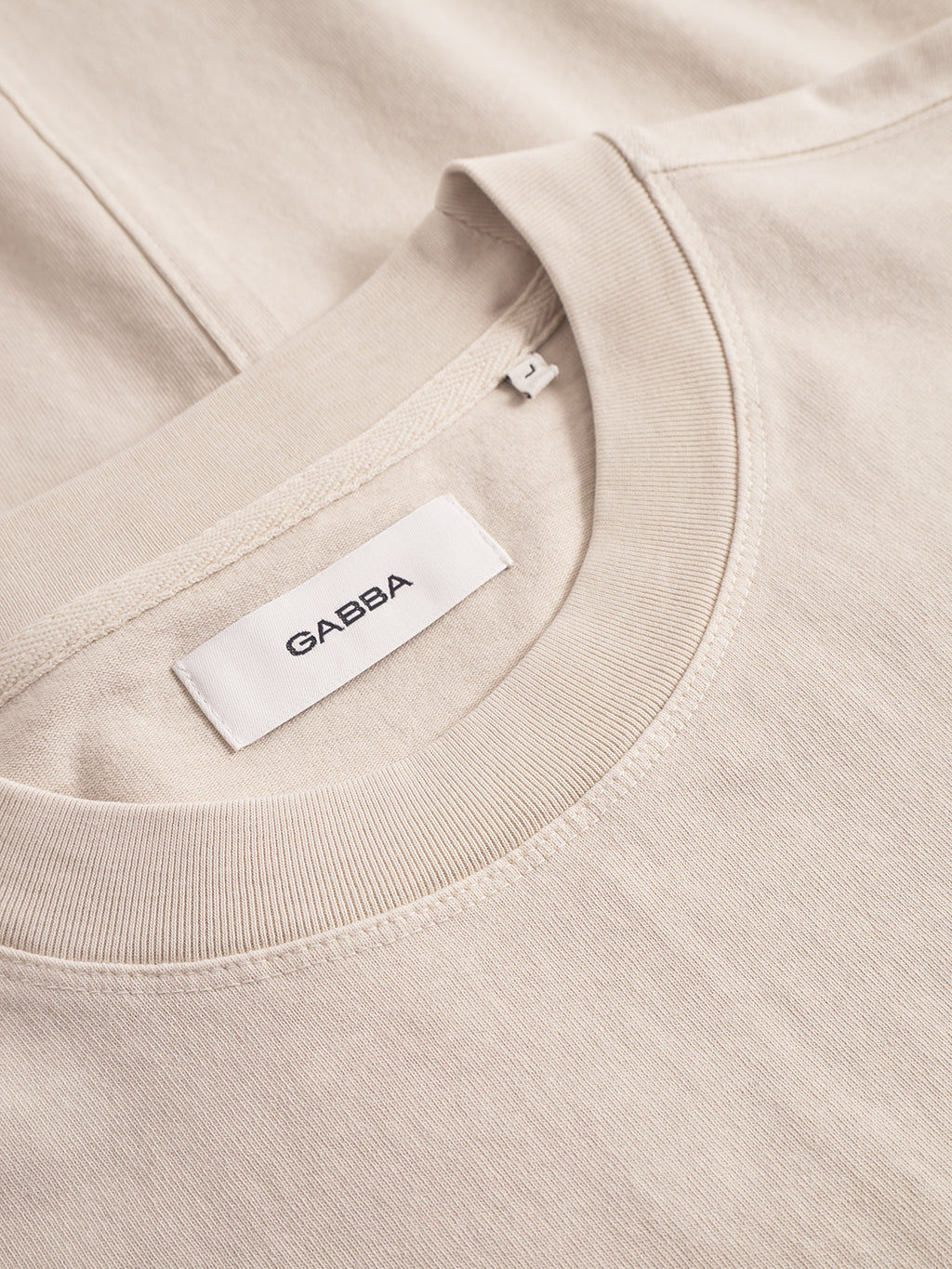A close-up of the Nigel Wix Boxy Hour SS T-shirt in White Almond shows soft, garment-dyed cotton with neat stitching and a white collar label featuring the Gabba brand name in black text.