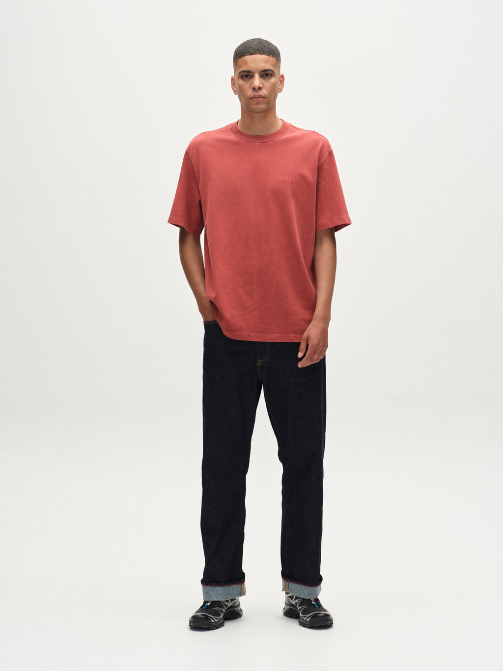 A man stands facing forward against a plain white background, wearing the Gabba Nigel Wix Boxy Hour SS tee in Red Russet. His hands are in his pockets, paired with dark cuffed jeans and black socks featuring a white pattern.