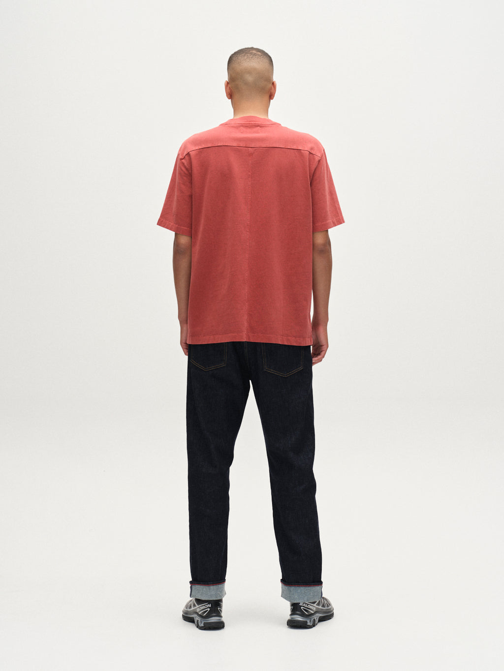 A person with short hair stands facing away, wearing the Gabba Nigel Wix Boxy Hour SS tee in Red Russet, dark cuffed jeans, and gray sneakers against a plain light background.