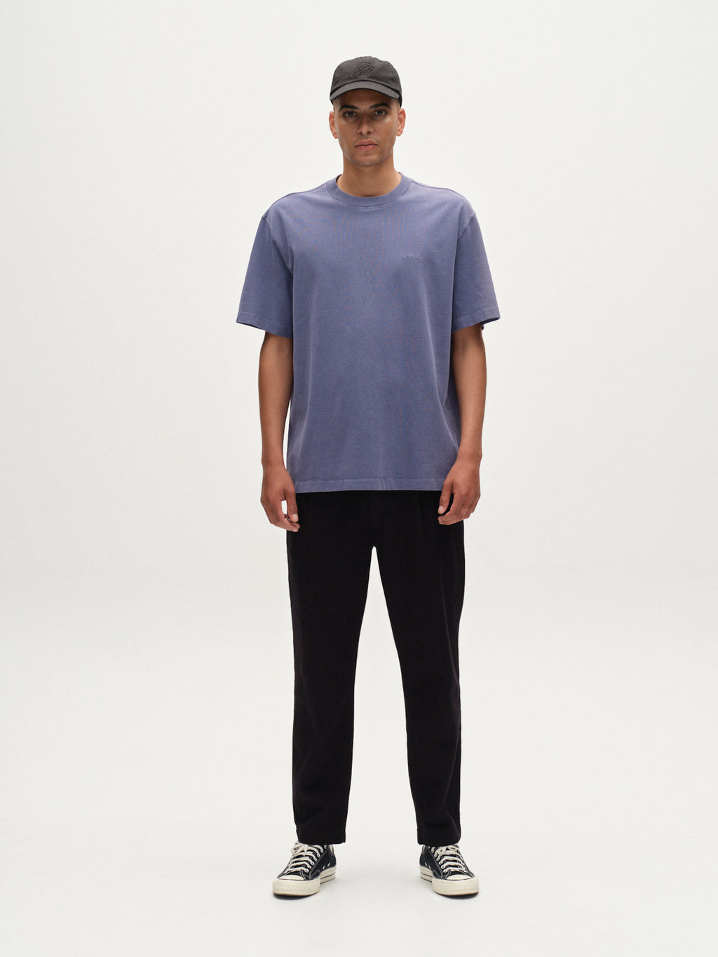 A person stands facing forward against a white background, wearing a black cap, Gabbas Nigel Wix Boxy Hour SS t-shirt in Vintage Indigo, black pants, and black-and-white sneakers.