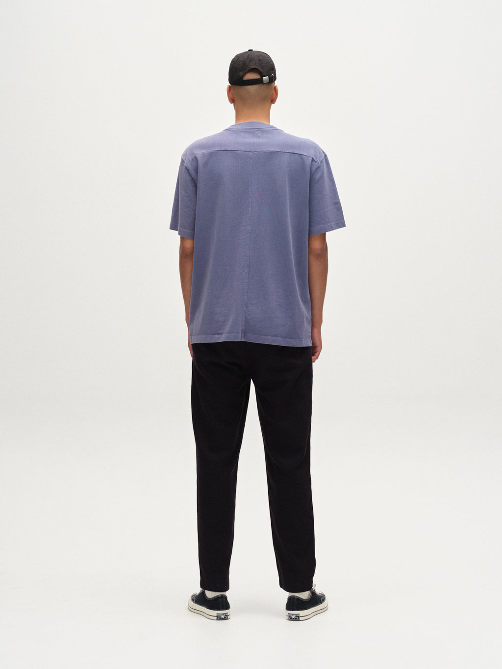 A man faces away from the camera against a white background, wearing a black cap, black pants, black sneakers with white soles, and the Gabba Nigel Wix Boxy Hour SS t-shirt in vintage indigo.