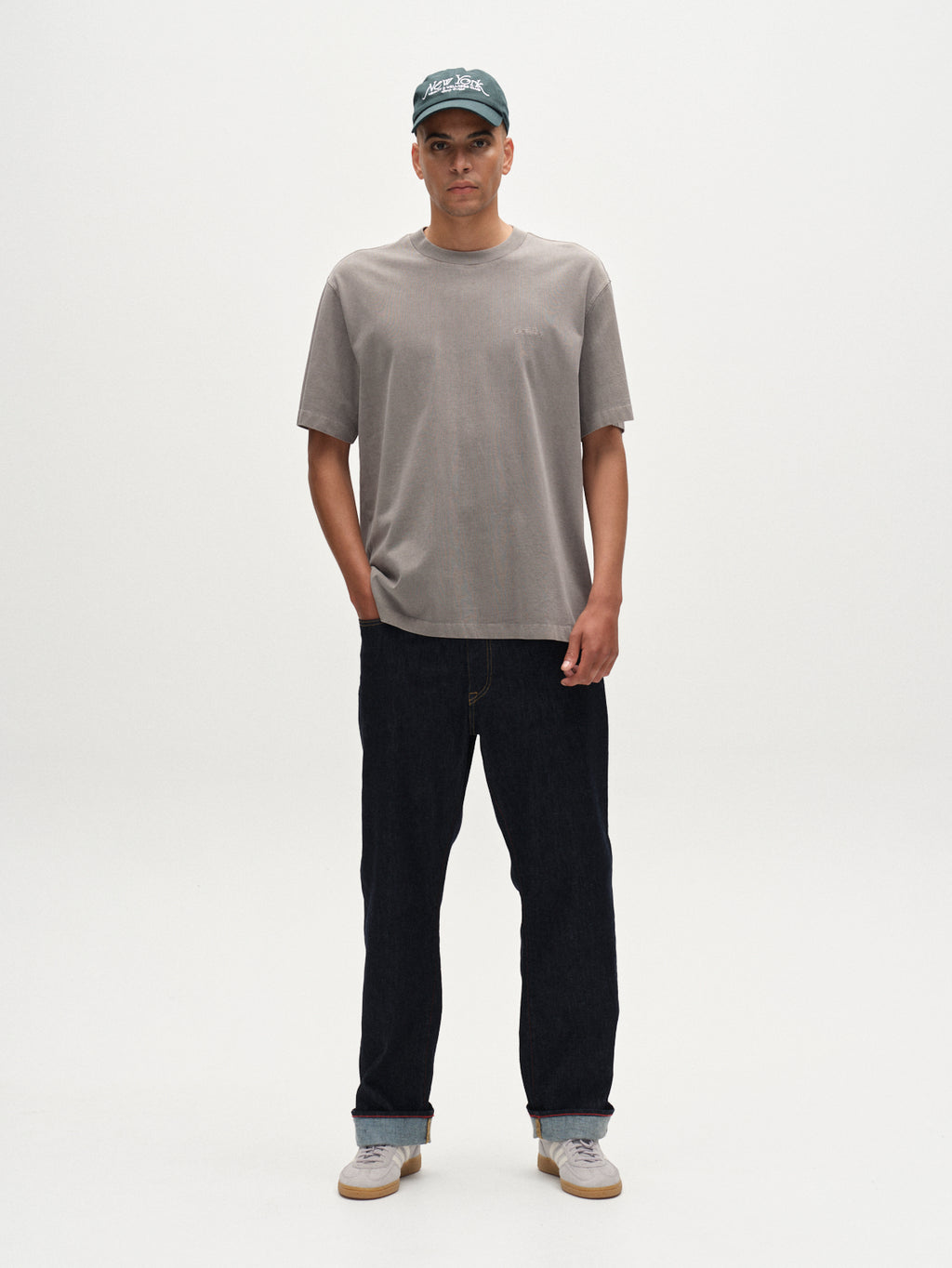 A man stands against a white background wearing the Gabba Nigel Wix Boxy Hour SS t-shirt in Dust Sage Green, paired with dark jeans, gray sneakers, and a green baseball cap. His hands are in his pockets and he faces forward neutrally.