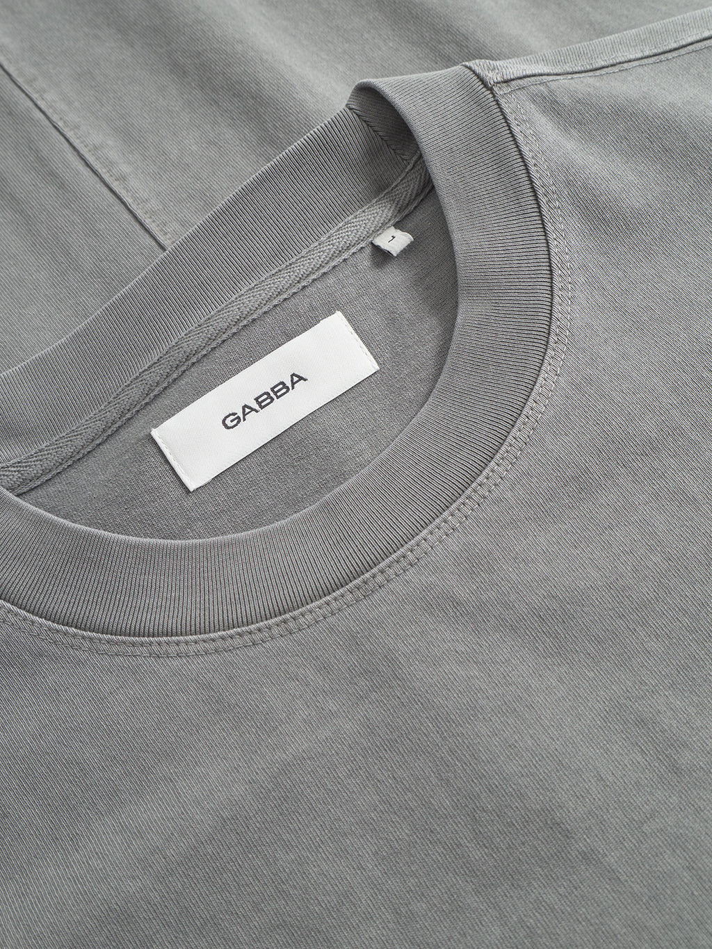 A close-up of a sage green boxy-fit cotton t-shirt, the Nigel Wix Boxy Hour SS by Gabba, with a visible white Gabba label sewn inside the collar.