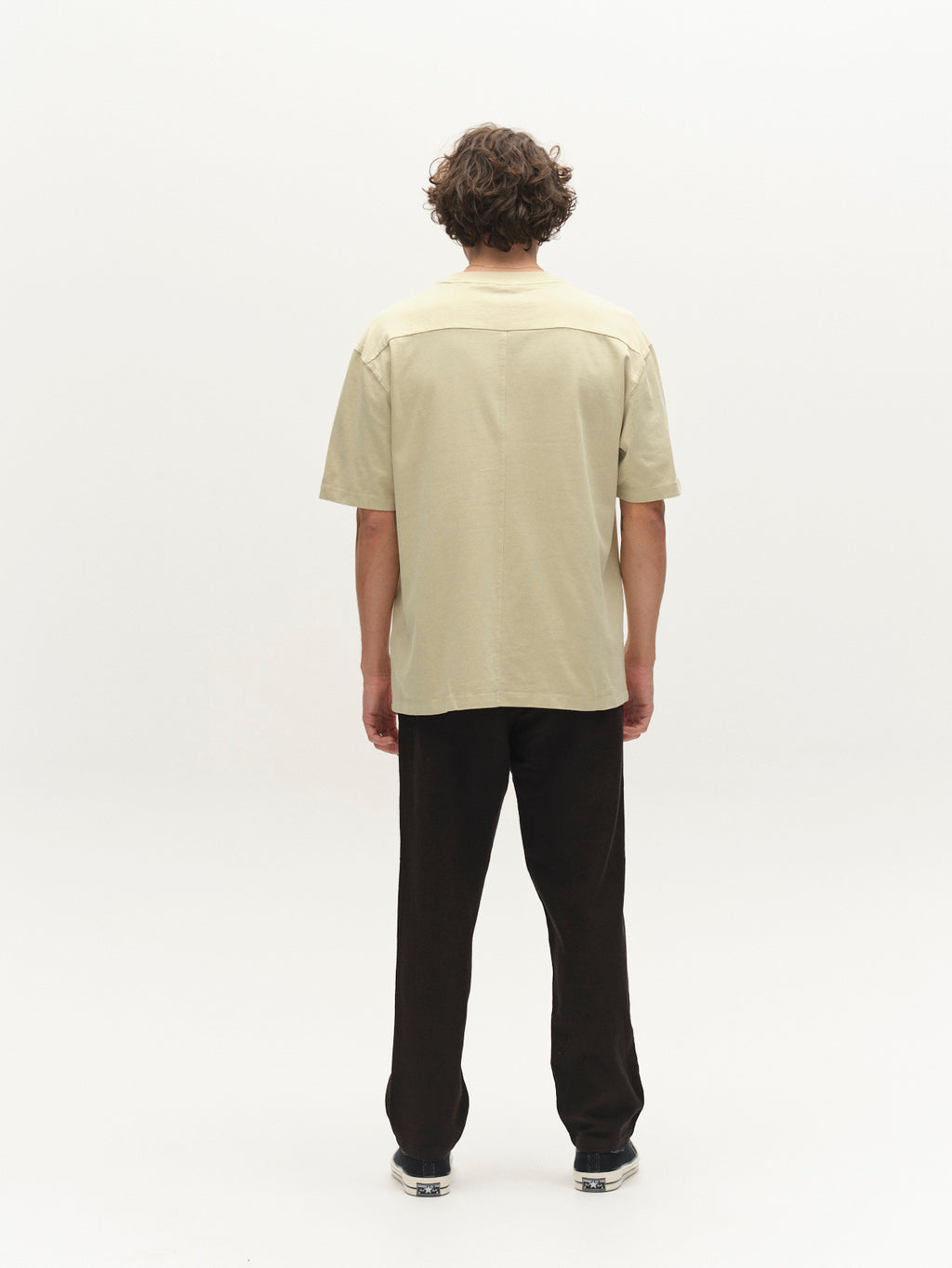 A person with curly hair faces away from the camera, wearing the Gabba GABnigel Wix Boxy Hour SS t-shirt in Alfa Green, black pants, and sneakers, set against a plain white background.