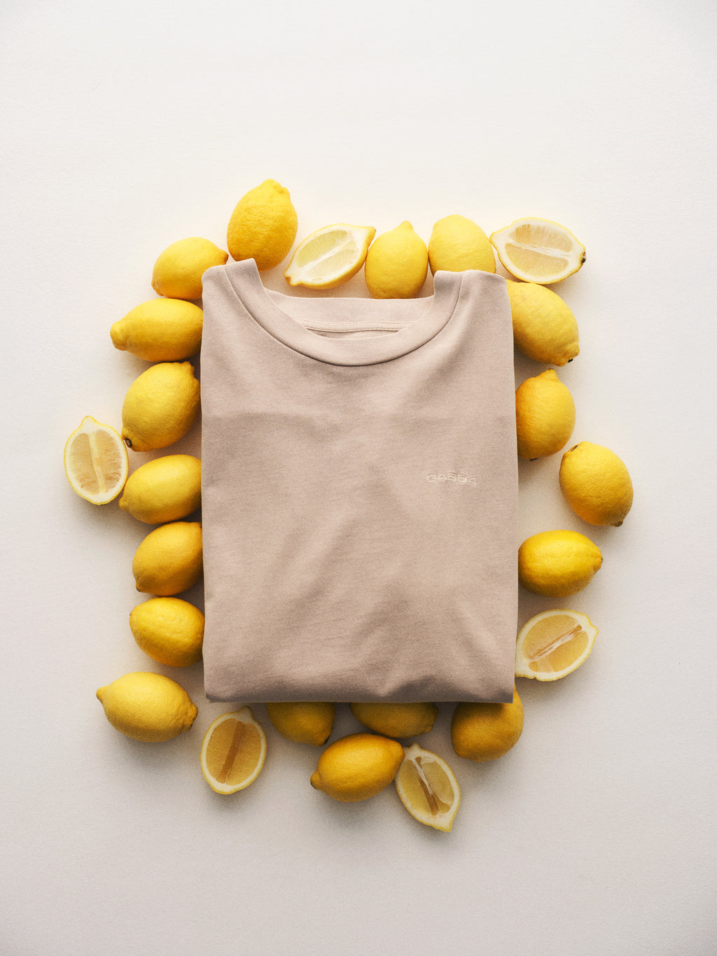 The Gabba GABnigel Wix Boxy Hour SS - Sand Cashmere t-shirt, crafted from heavyweight cotton, lies folded on a white surface with whole and halved yellow lemons arranged in an oval around it, evoking effortless style and comfort.