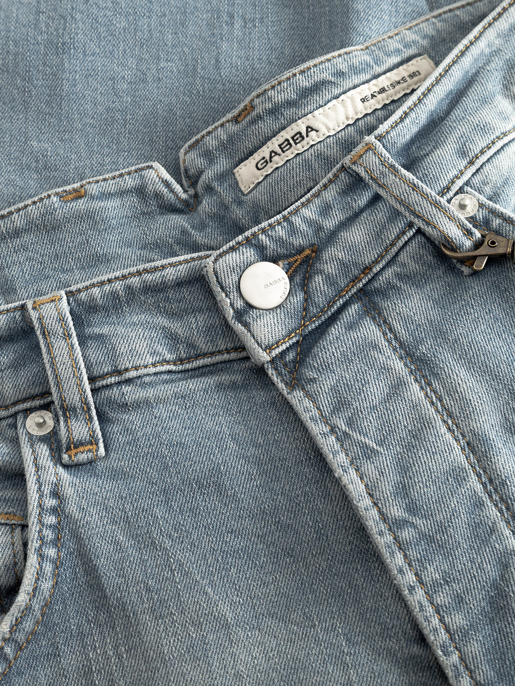 Close-up of Gabba GABalex Cros Cropped light blue denim jeans with visible seams, metal button, belt loops, and a white inner waistband label. These relaxed fit jeans are laid flat on matching cross-hatch fabric denim.