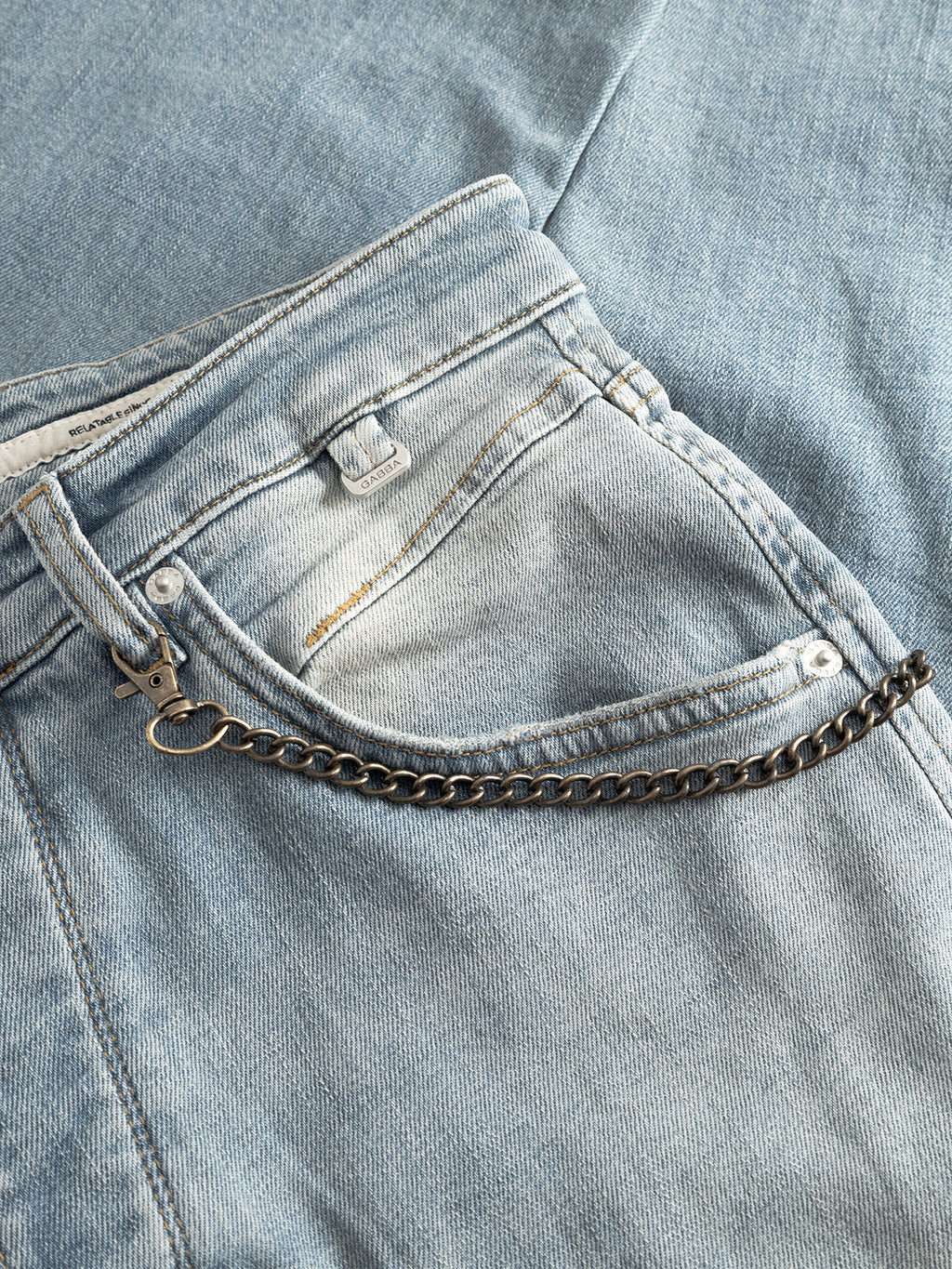 A close-up of the Gabba GABalex Cros Cropped relaxed fit jeans in light blue denim, with a metal chain detail on the front pocket, displayed on matching fabric.
