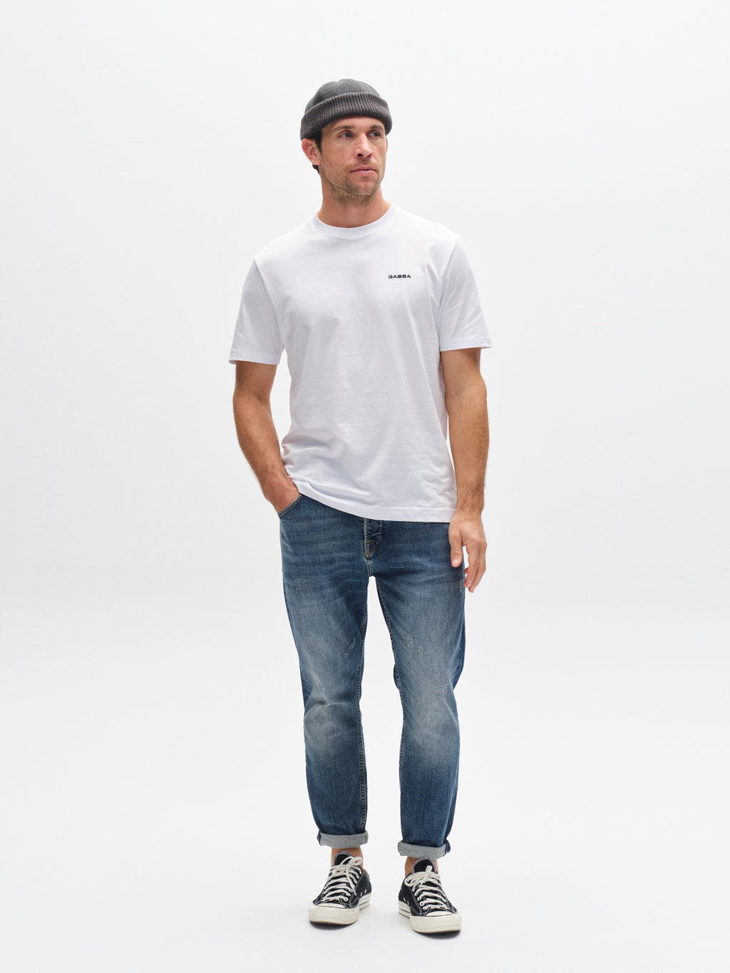 A man wearing a gray beanie, plain white t-shirt, Gabba’s GABalex Cros Cropped relaxed fit jeans in mid blue denim, and black-and-white sneakers stands against a plain white background.