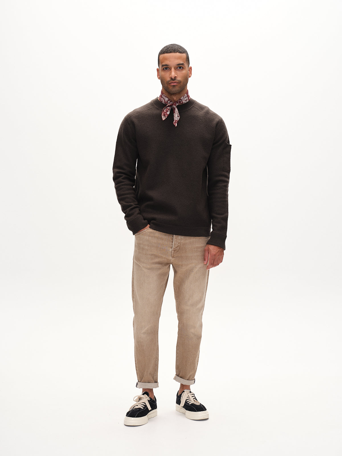 A man faces forward with arms relaxed at his sides, wearing Gabbas Alex Earth - Sand relaxed-fit beige denim jeans, a dark sweater, black sneakers, and a red patterned bandana against a plain white background.