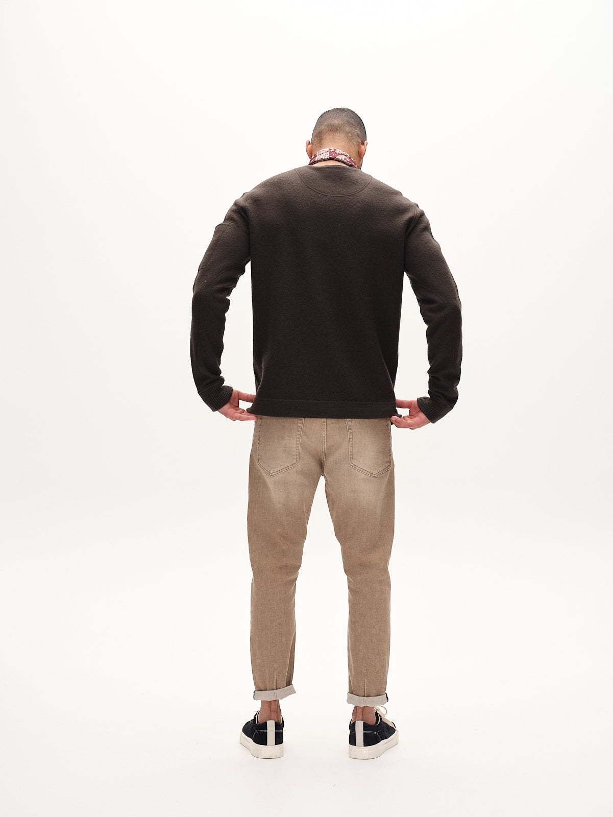 A man stands with his back to the camera against a white background, wearing Gabbas Alex Earth - Sand cuffed jeans, a dark brown sweater, black and white sneakers, and a red scarf. His hands are on his hips.