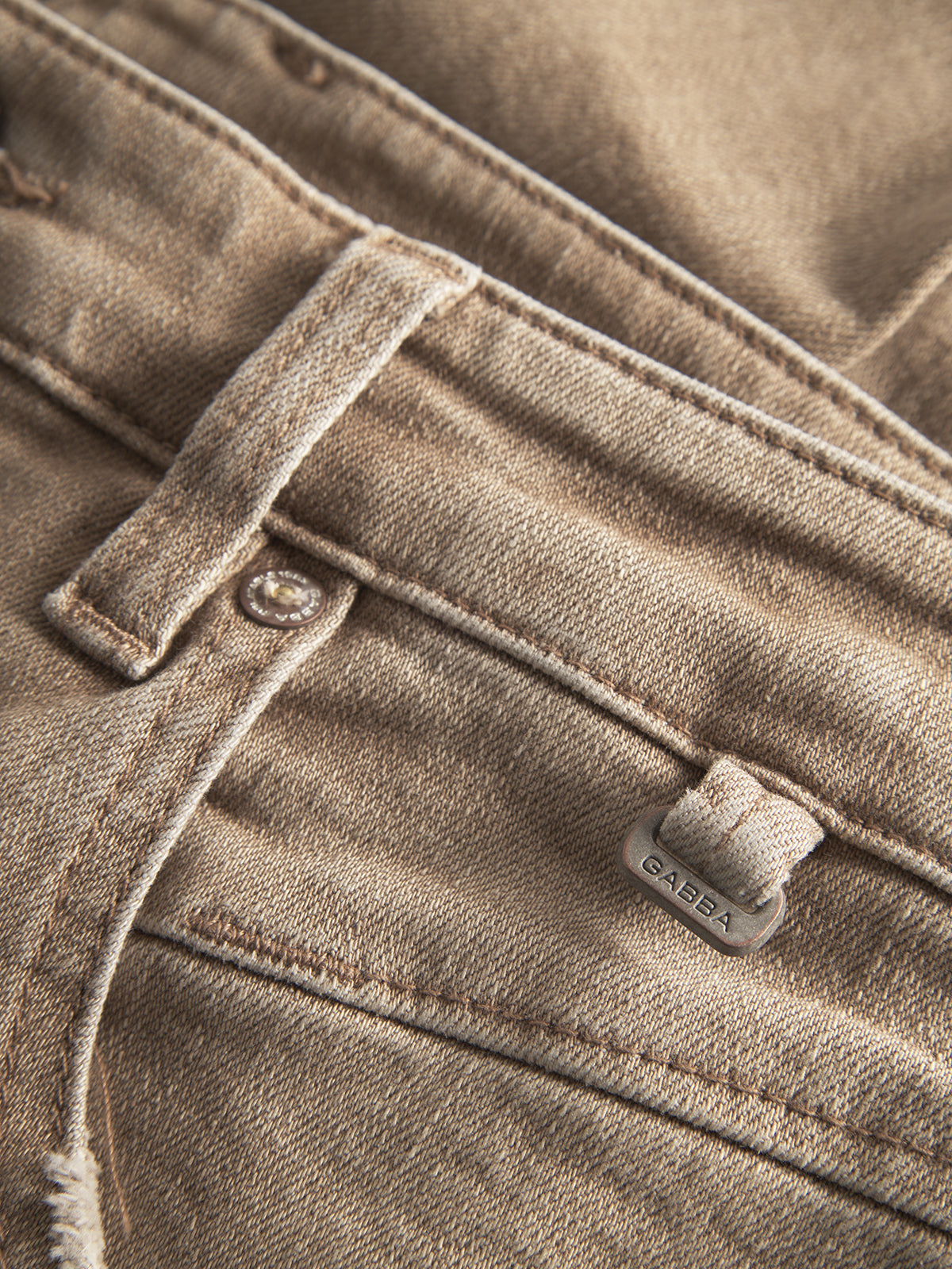Close-up of Alex Earth - Sand denim jeans by Gabba, featuring a belt loop, waistband, pocket, and a metal tag with the brand name. The relaxed-fit jeans show detailed stitching and fabric texture in a sand-toned finish.