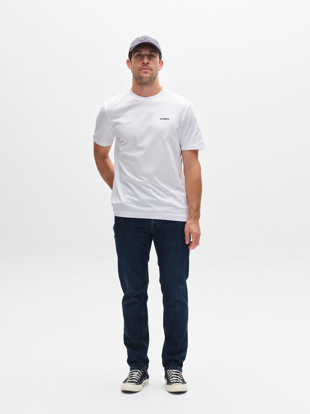A man stands facing forward against a white background, wearing a gray cap, white t-shirt, Gabba GABmarc Mego - Dark Blue Denim jeans, and black-and-white sneakers. His left arm is behind his back and his right arm hangs at his side.