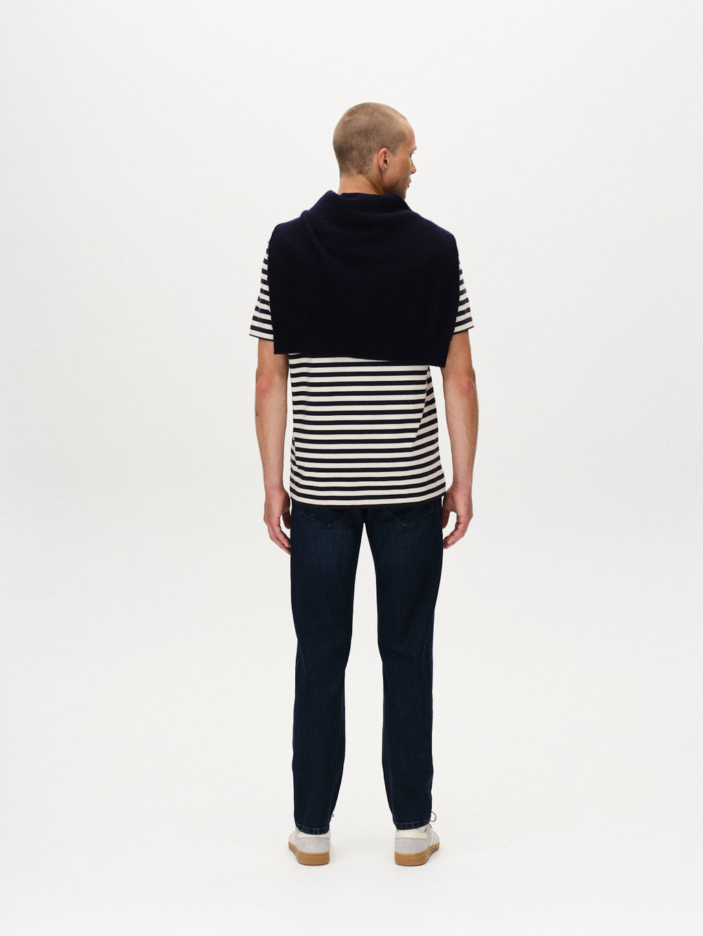 A man faces away from the camera, wearing a black and white striped T-shirt, Gabba Marc Mego Dark Blue Denim jeans with Wiser Wash technology, white sneakers, and a dark sweater over his shoulders. The background is plain white.