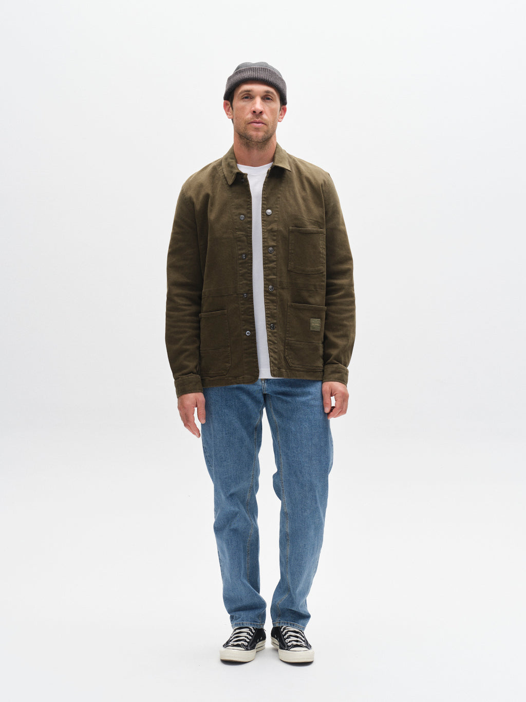 A man stands against a white background wearing Gabba’s GABmax Cros - Lt. Blue Denim jeans, styled with an olive slim-fit jacket, light shirt, and black-and-white sneakers. He faces forward with a neutral look, highlighting a sharp, clean style.