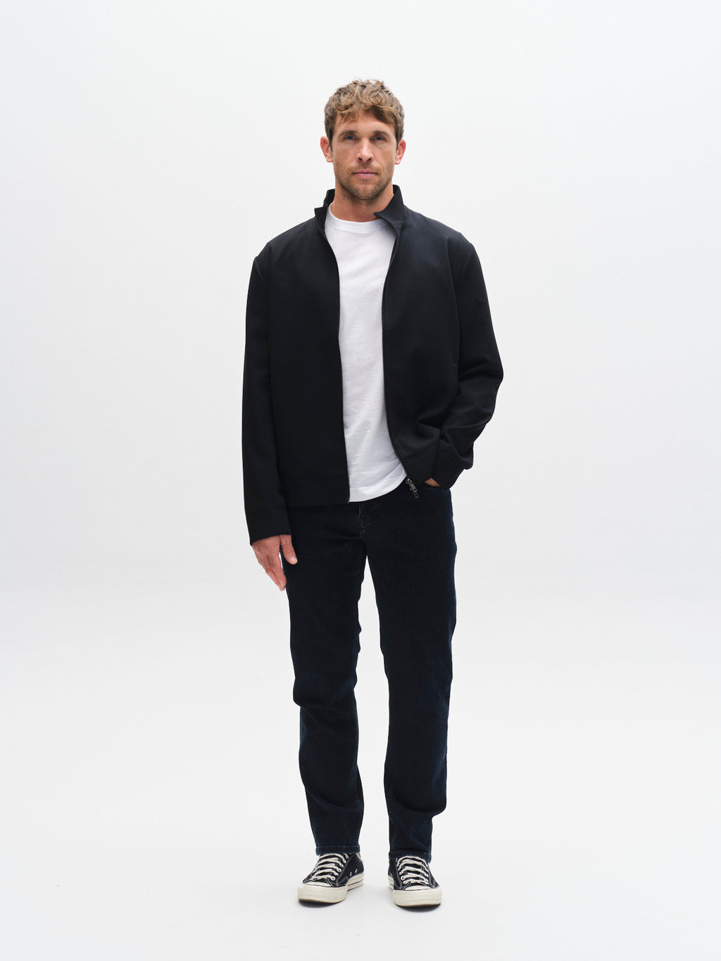 A man stands against a plain white background, facing forward with one hand in his pocket. He wears a black jacket over a white t-shirt, Gabba GABmarc Panter dark blue denim jeans, and black-and-white sneakers.