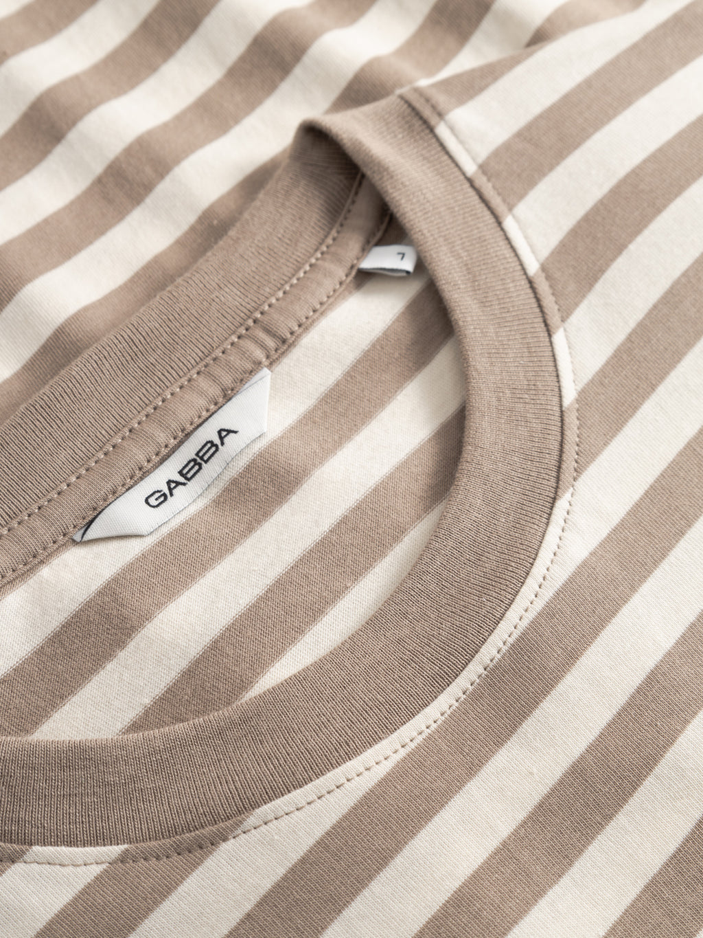 Close-up of Gabba’s GABdune Logo Stripes SS GOTS - Fungi, an organic cotton T-shirt in beige and off-white stripes with the Gabba label by the collar. The fabric looks soft and the neckline features neat stitching.
