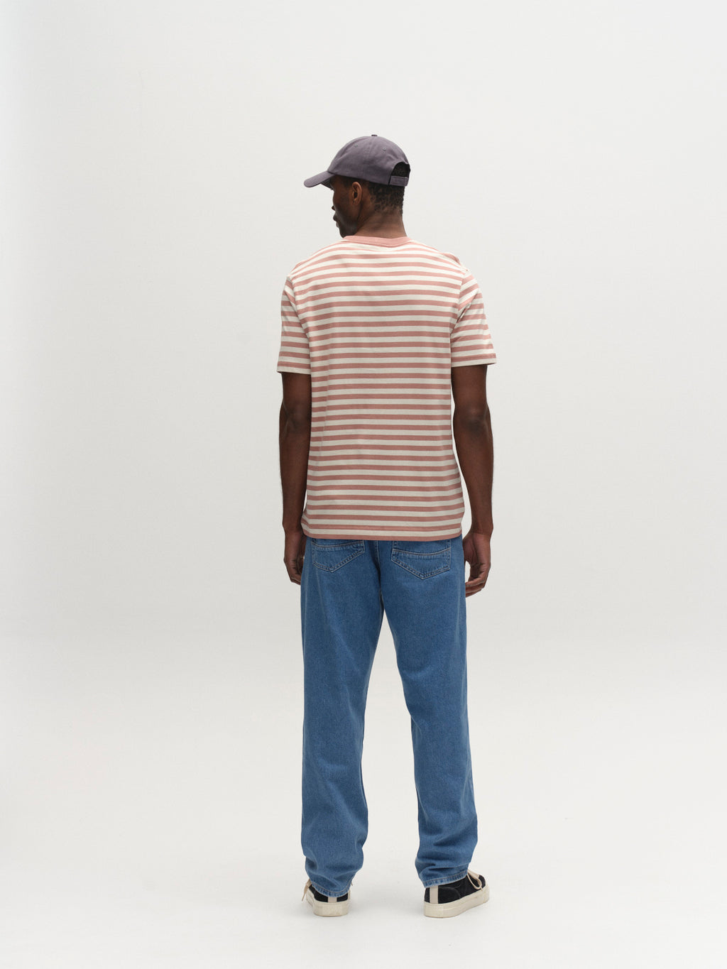 A man faces away from the camera, wearing a Gabba GABdune Logo Stripes SS GOTS t-shirt in Wood Pink, blue jeans, a gray cap, and black-and-white sneakers, set against a plain white background.