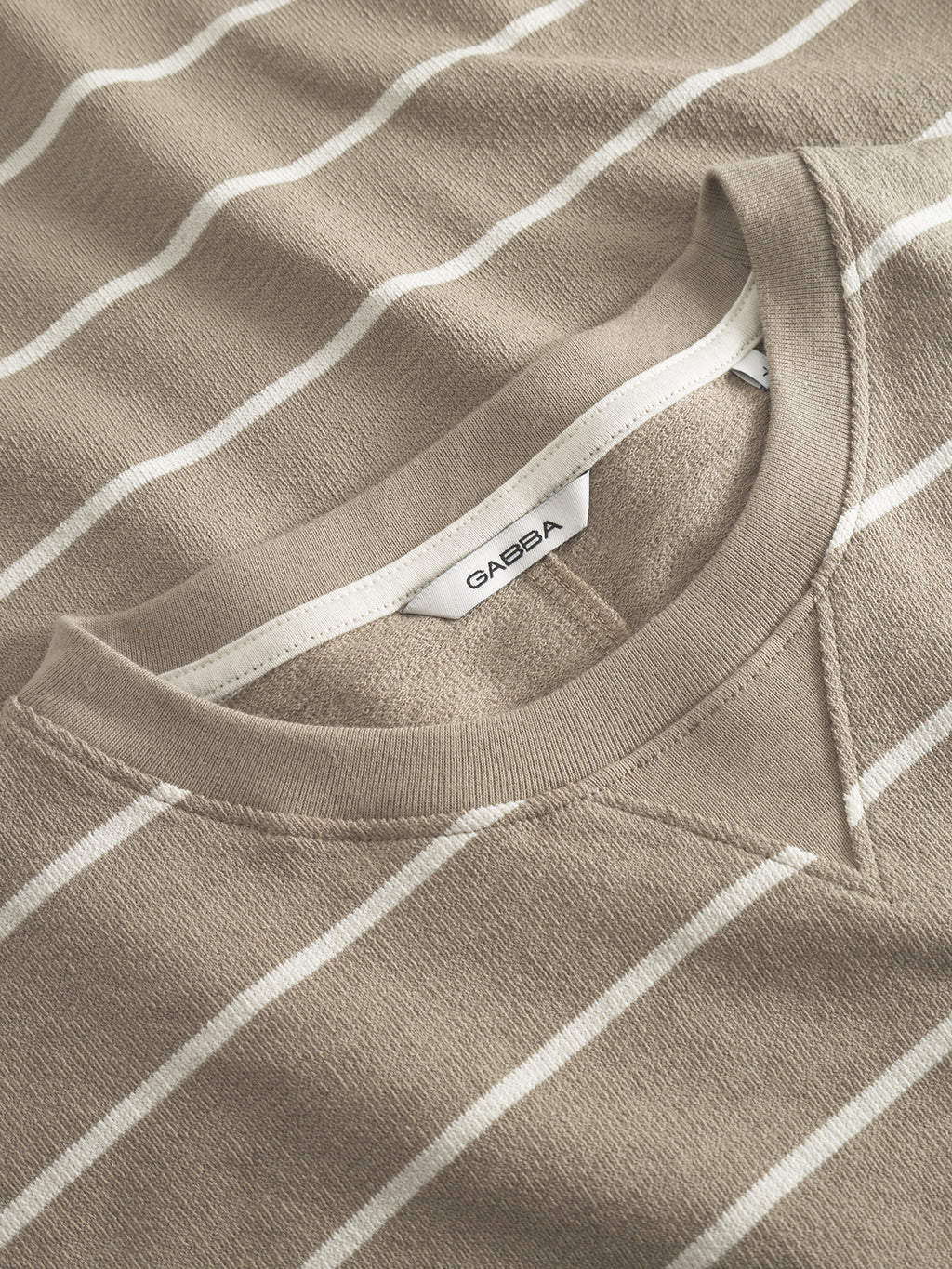 Close-up of Gabbas Dune Crepe Stripes SS GOTS in brown stripe, made from GOTS-certified organic cotton. Features a crew neckline with a visible white GAEBA label; fabric texture and stitching details are clear.