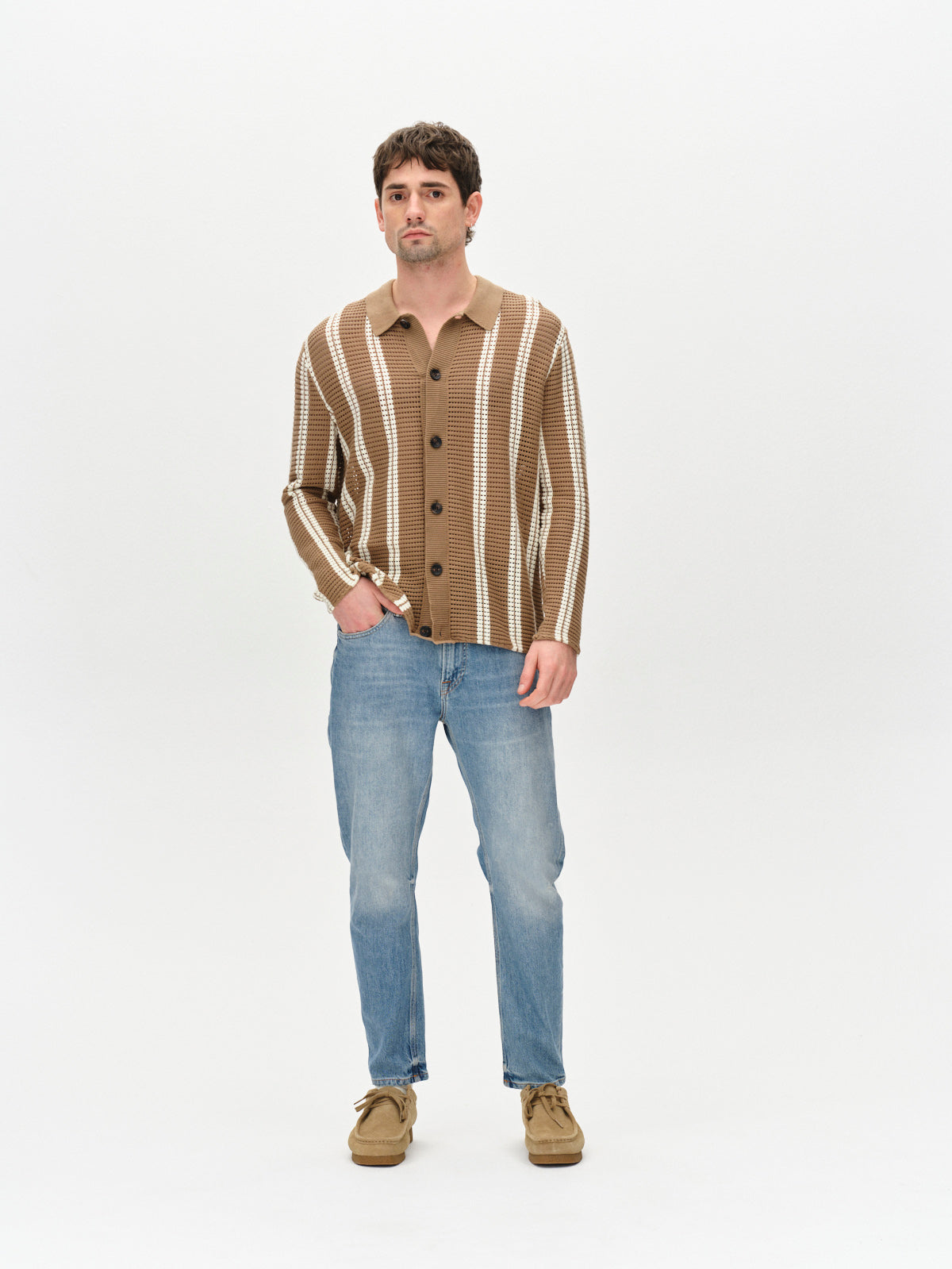 A man stands against a plain white background, effortlessly showcasing a casual stylish look. Hes wearing light blue Carl Astra - Mid Blue Denim jeans with a loose tapered fit by Gabba. His brown and white striped cardigan and brown shoes complement his short dark hair and neutral expression as he looks forward.