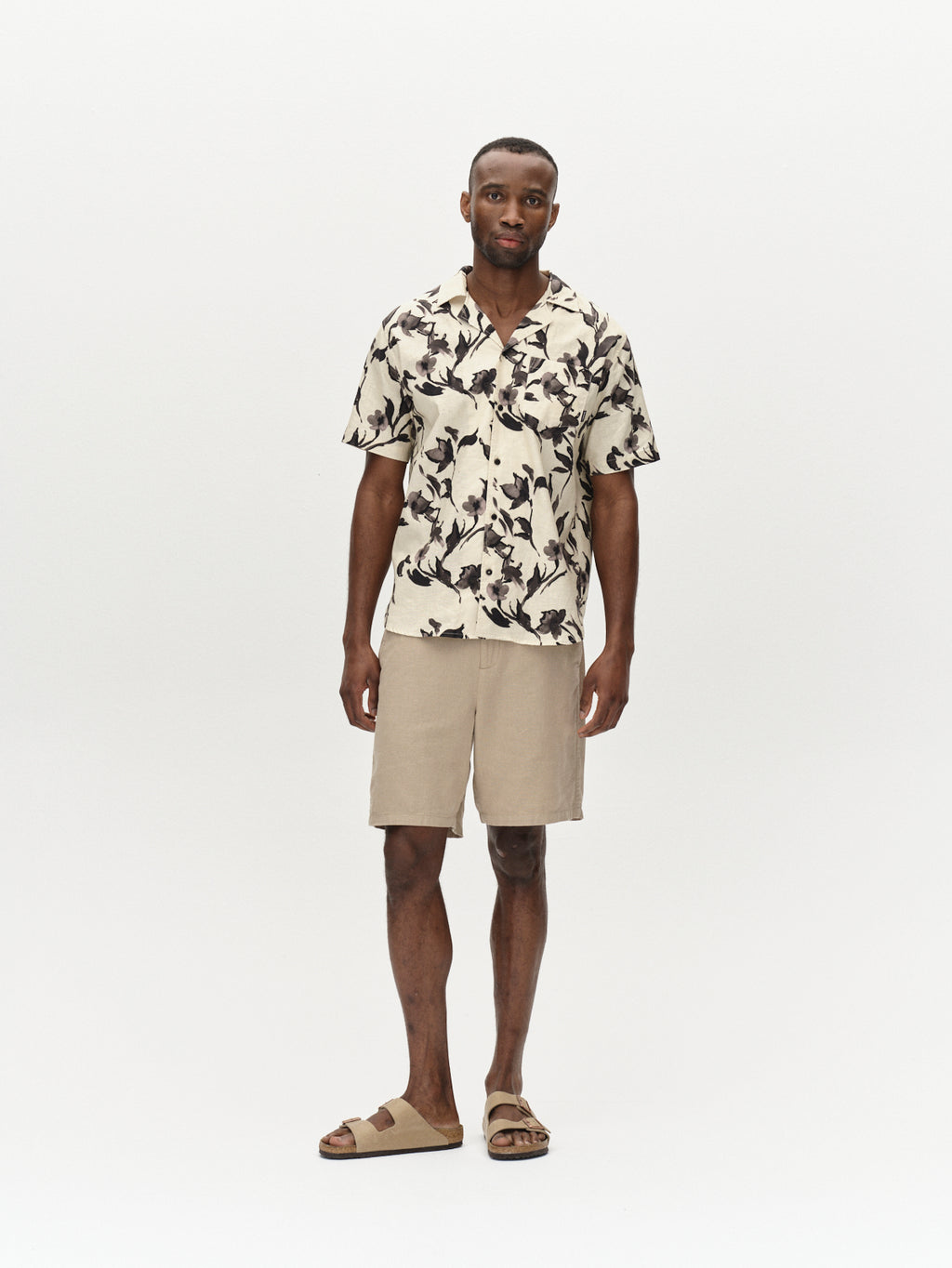 A man stands facing forward wearing Gabba's Sven Reg Resort Bloom SS - Meteorite shirt, beige shorts, and beige slide sandals against a plain white background.