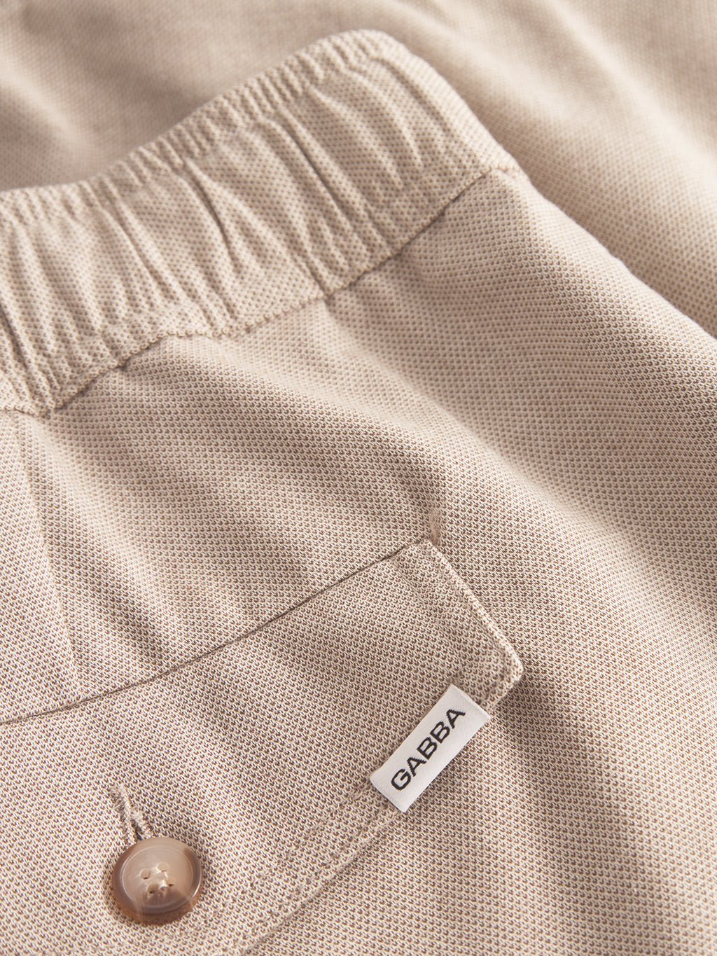 Close-up of Gabba Coz Belt Carre Shorts in sand: beige textured fabric, elastic waistband, back pocket with brown button, and a small white Gabba tag attached.